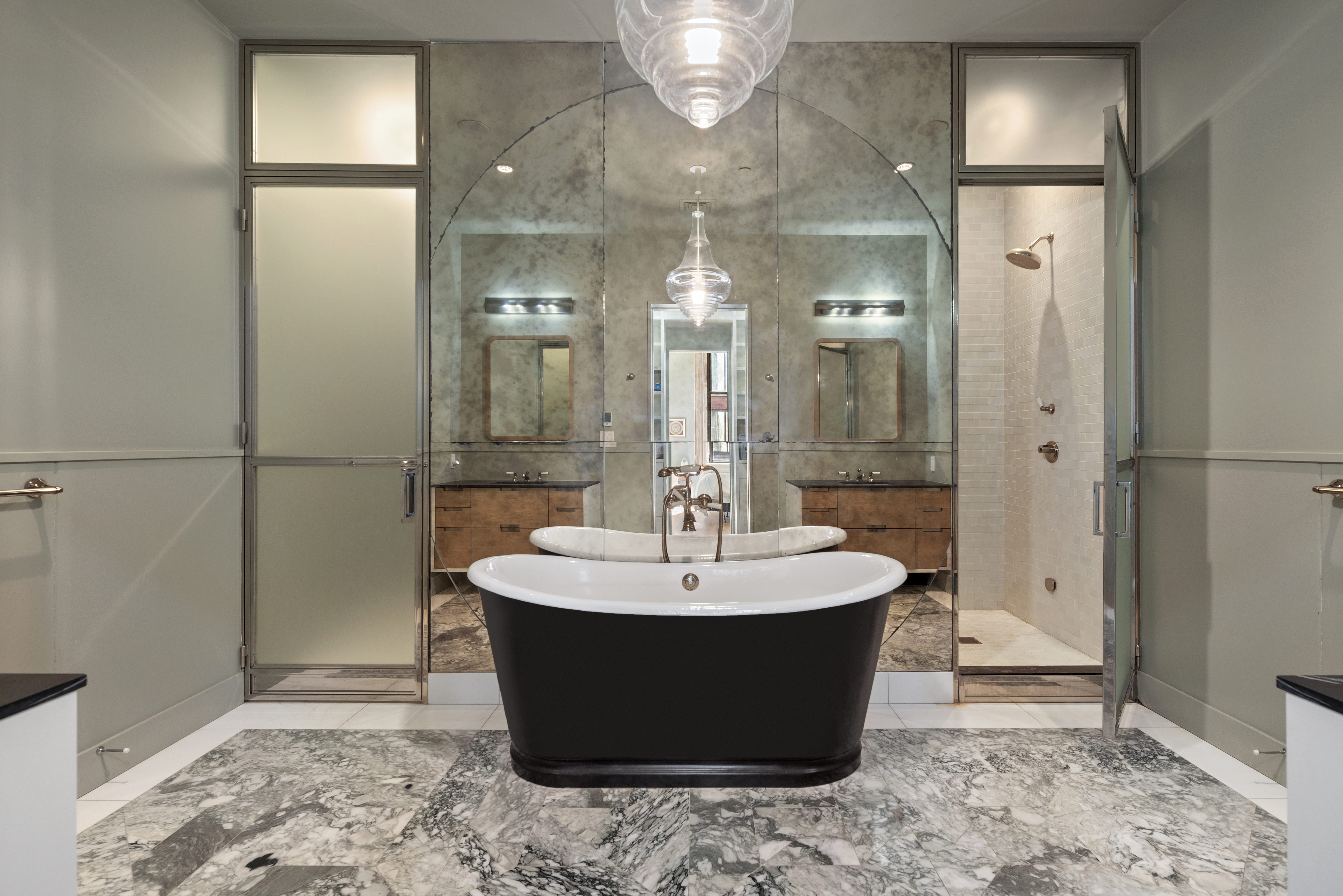 A luxurious bathroom with a black freestanding bathtub on a marble floor, glass doors leading to separate shower and toilet areas, and a large mirror reflecting a double-sink vanity with wooden cabinets and modern lighting.
