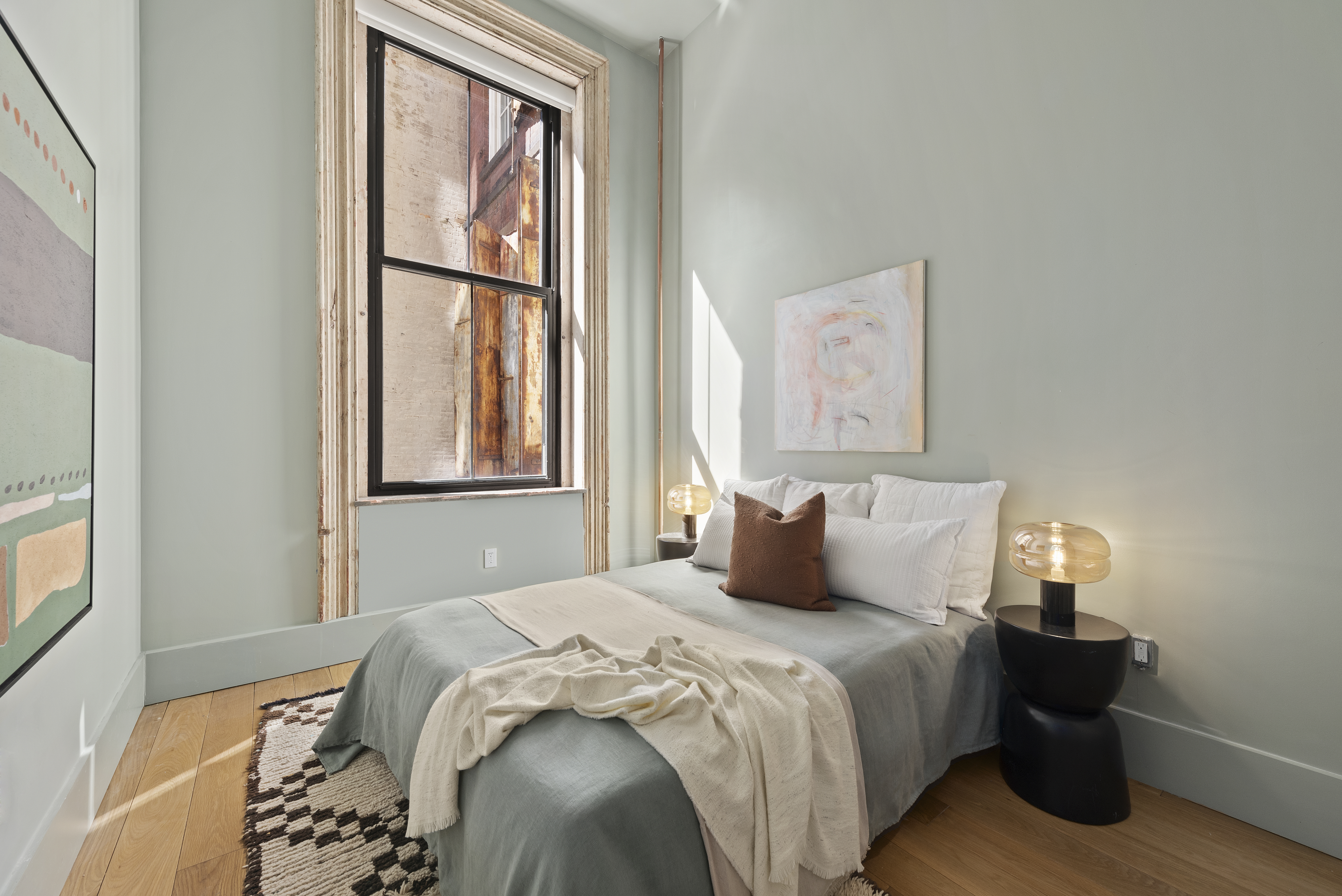 A well-lit bedroom with light green walls, wooden flooring, a bed with gray bedding and white and brown accent pillows, and two black nightstands with modern lamps beside a large window.