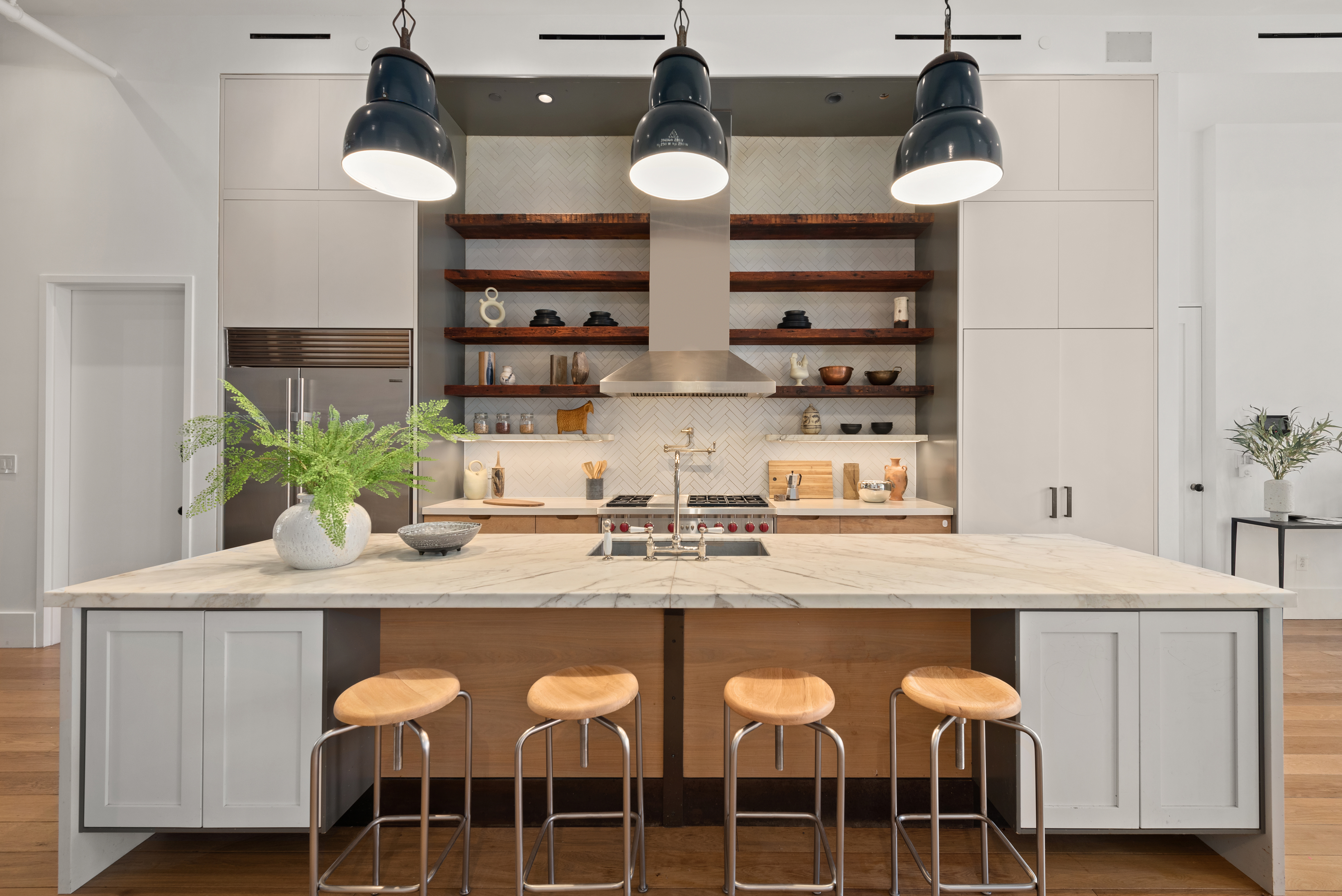 Modern kitchen with a large marble island, four wooden stools, three black pendant lights, open shelves with kitchen items, a stainless steel refrigerator, and white cabinetry.