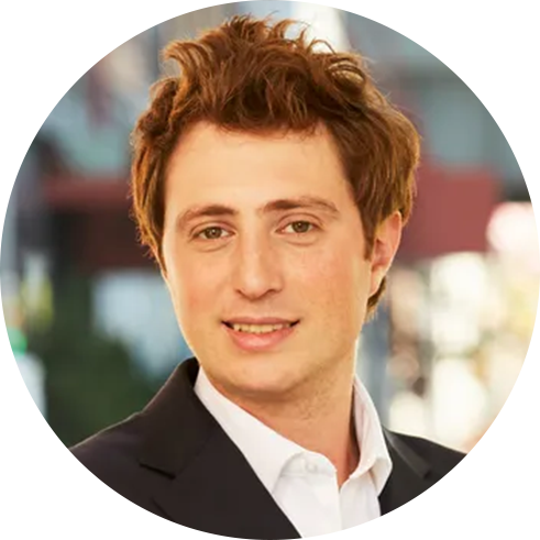 A person with short, wavy brown hair wearing a white collared shirt and a dark suit jacket.