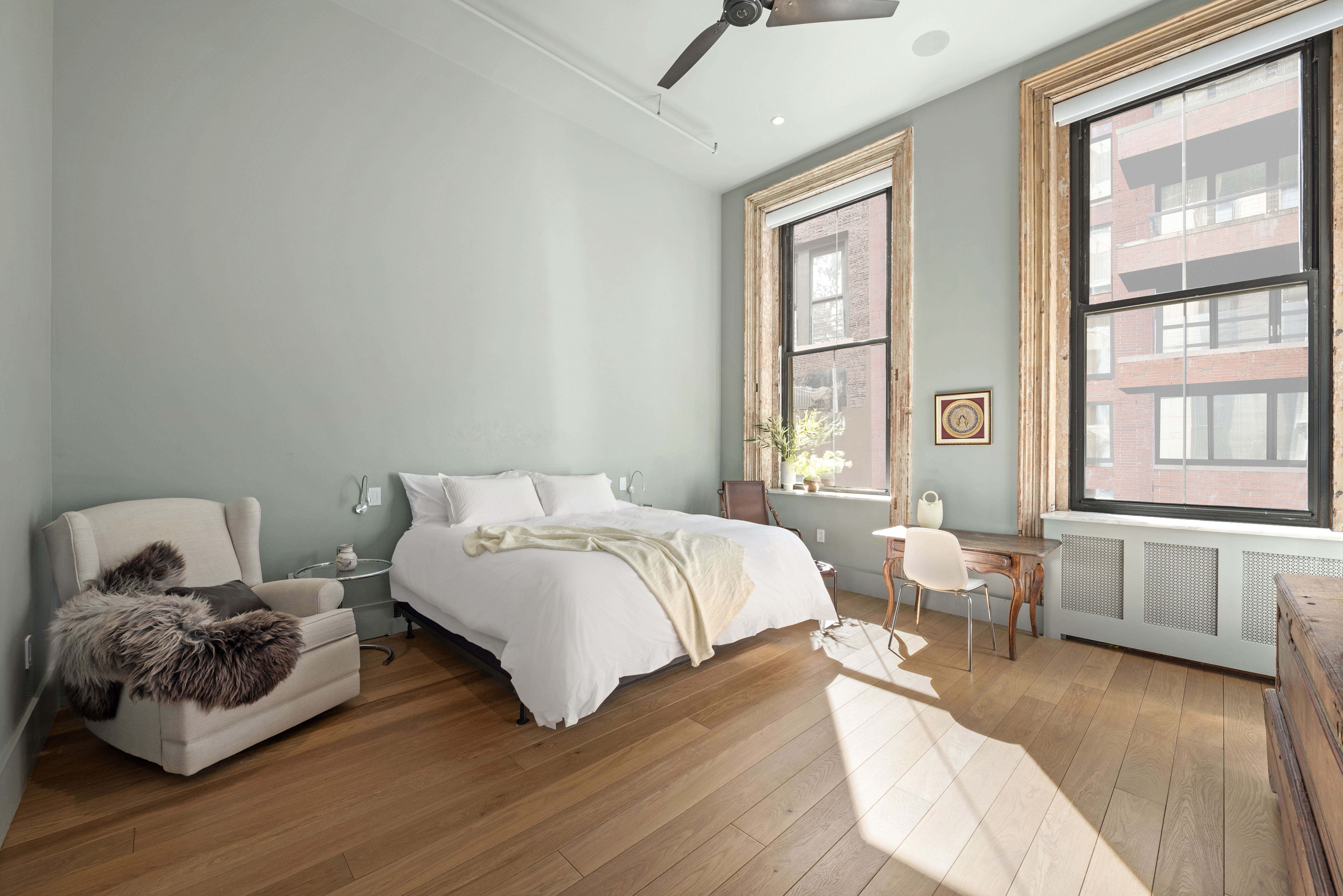 A spacious, well-lit bedroom with light green walls, a large bed with white bedding, a beige armchair with a fur throw, two large windows, a wooden desk and chair, and hardwood flooring.