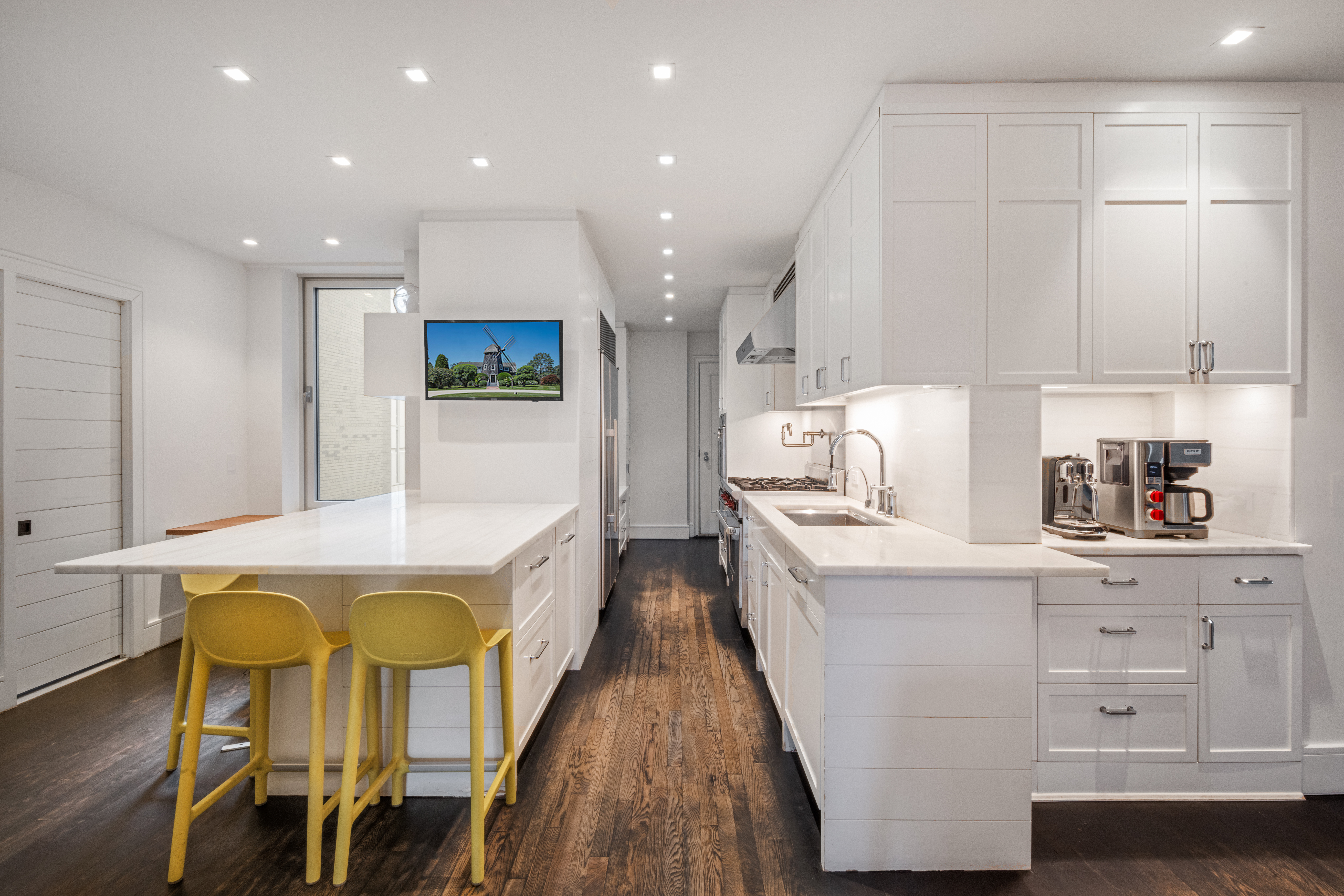 A modern kitchen with white cabinets, a large island with two yellow stools, stainless steel appliances, dark wooden flooring, recessed ceiling lights, and a small wall-mounted TV above the island.
