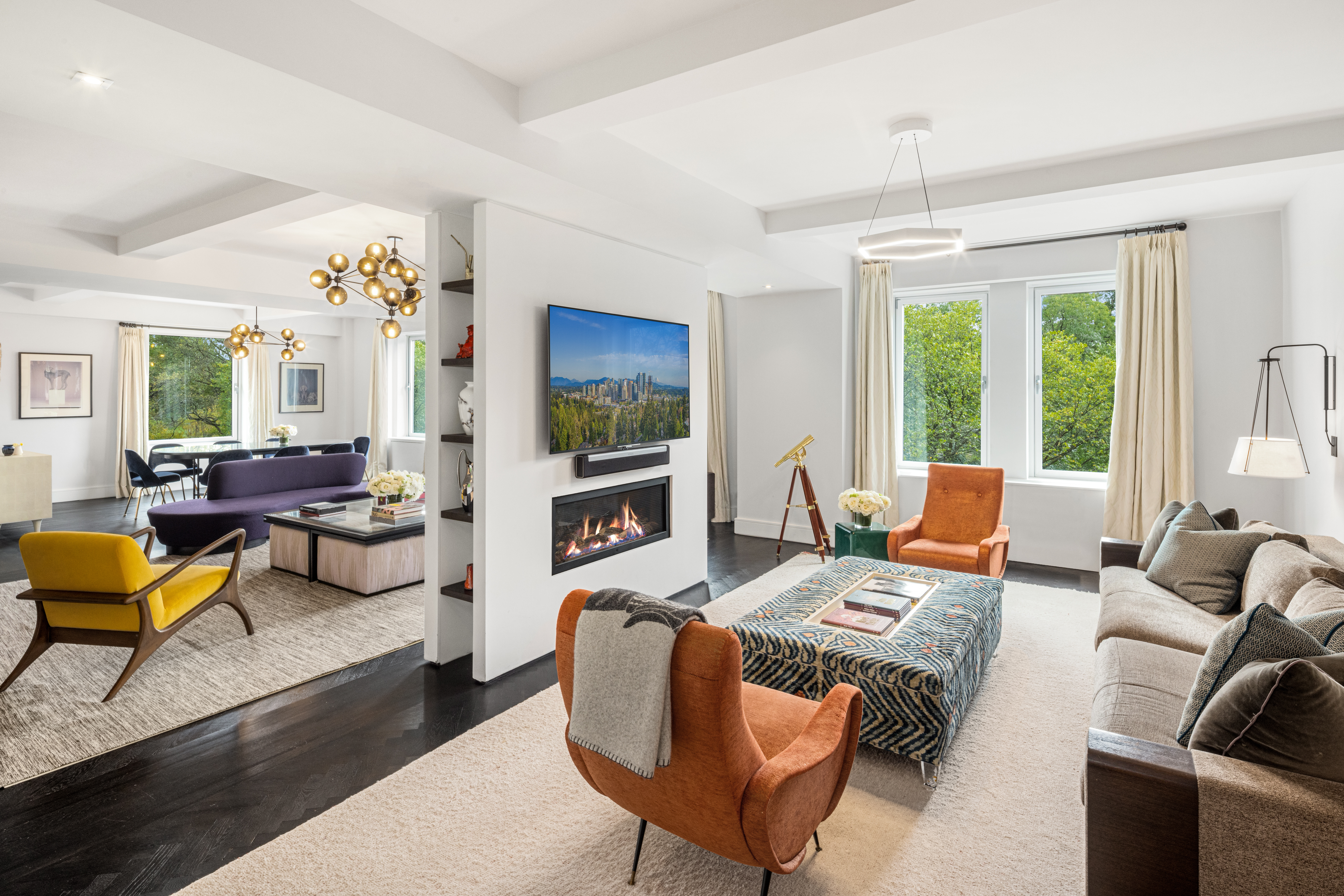 A modern living room with a fireplace, wall-mounted TV, large windows showing greenery, orange and yellow chairs, a gray sofa, and a patterned ottoman; a dining area with purple chairs is visible in the background.