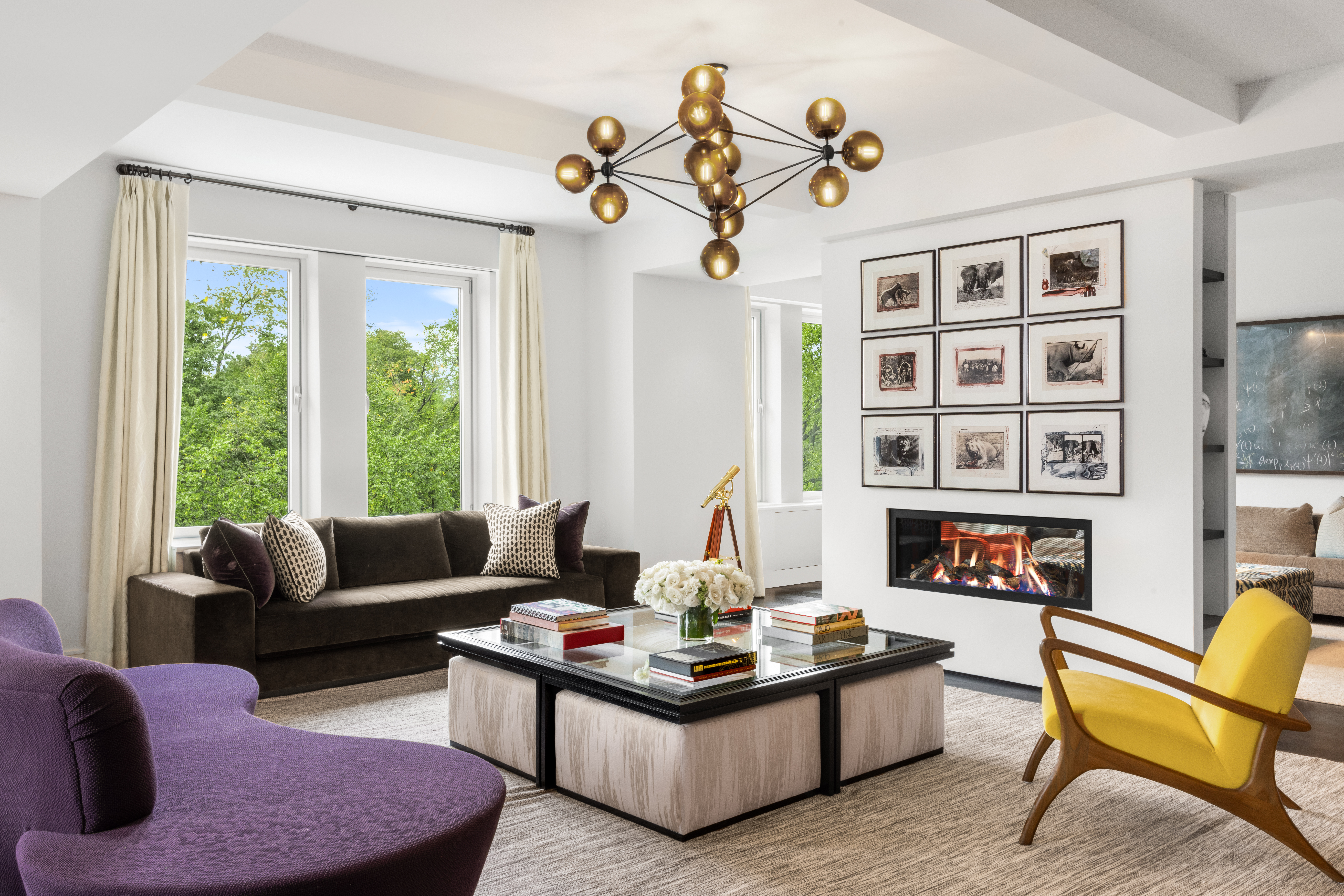 A modern living room with a large window, dark brown sofa, purple chaise lounge, yellow armchair, central coffee table with books and flowers, a fireplace with framed pictures above it, and an artistic chandelier hanging from the ceiling.
