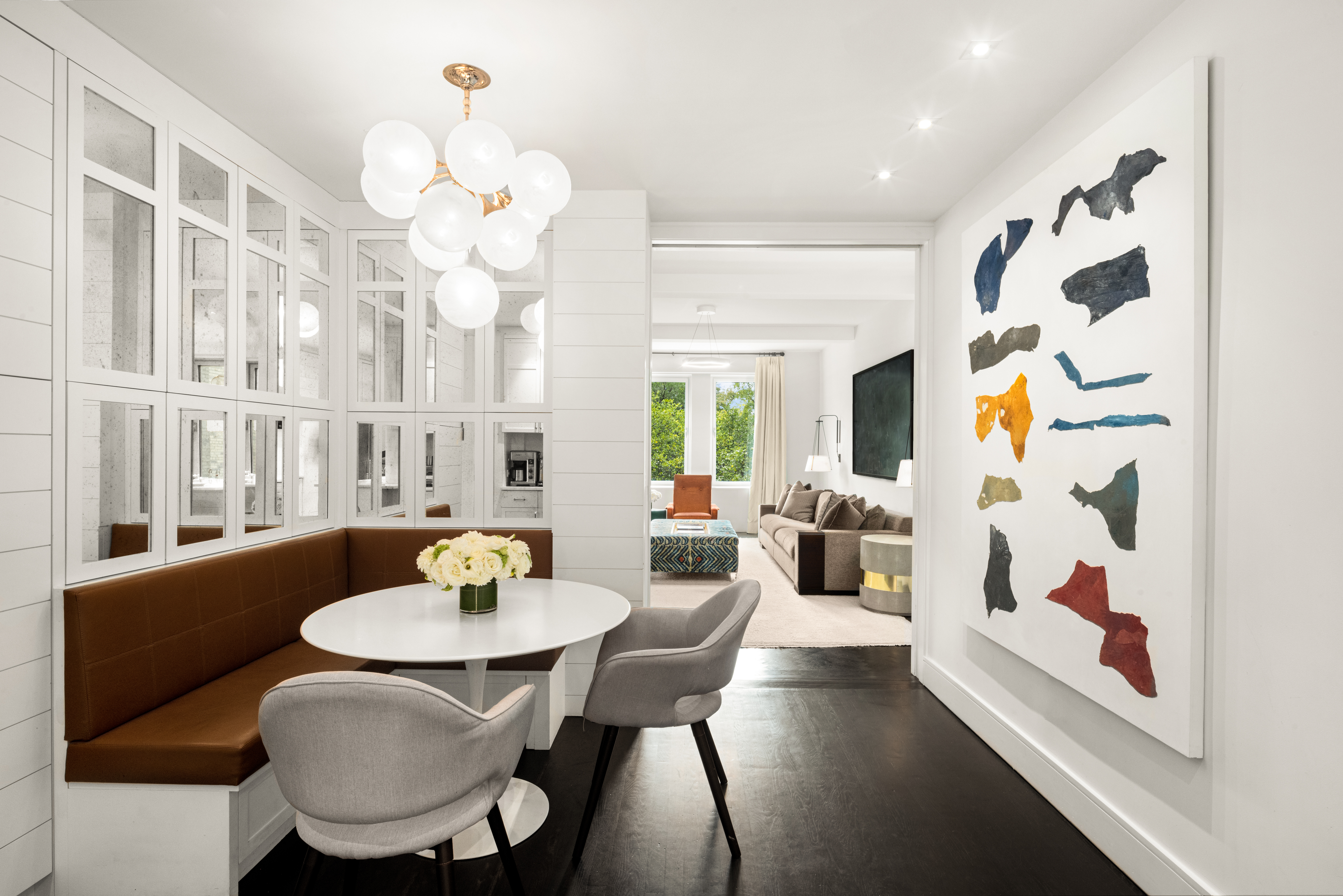 A modern dining area with a round white table, brown cushioned bench, and two gray chairs, lit by a chandelier with spherical lights; mirrored wall panels and a large abstract painting decorate the space, with an open view into a contemporary living room.