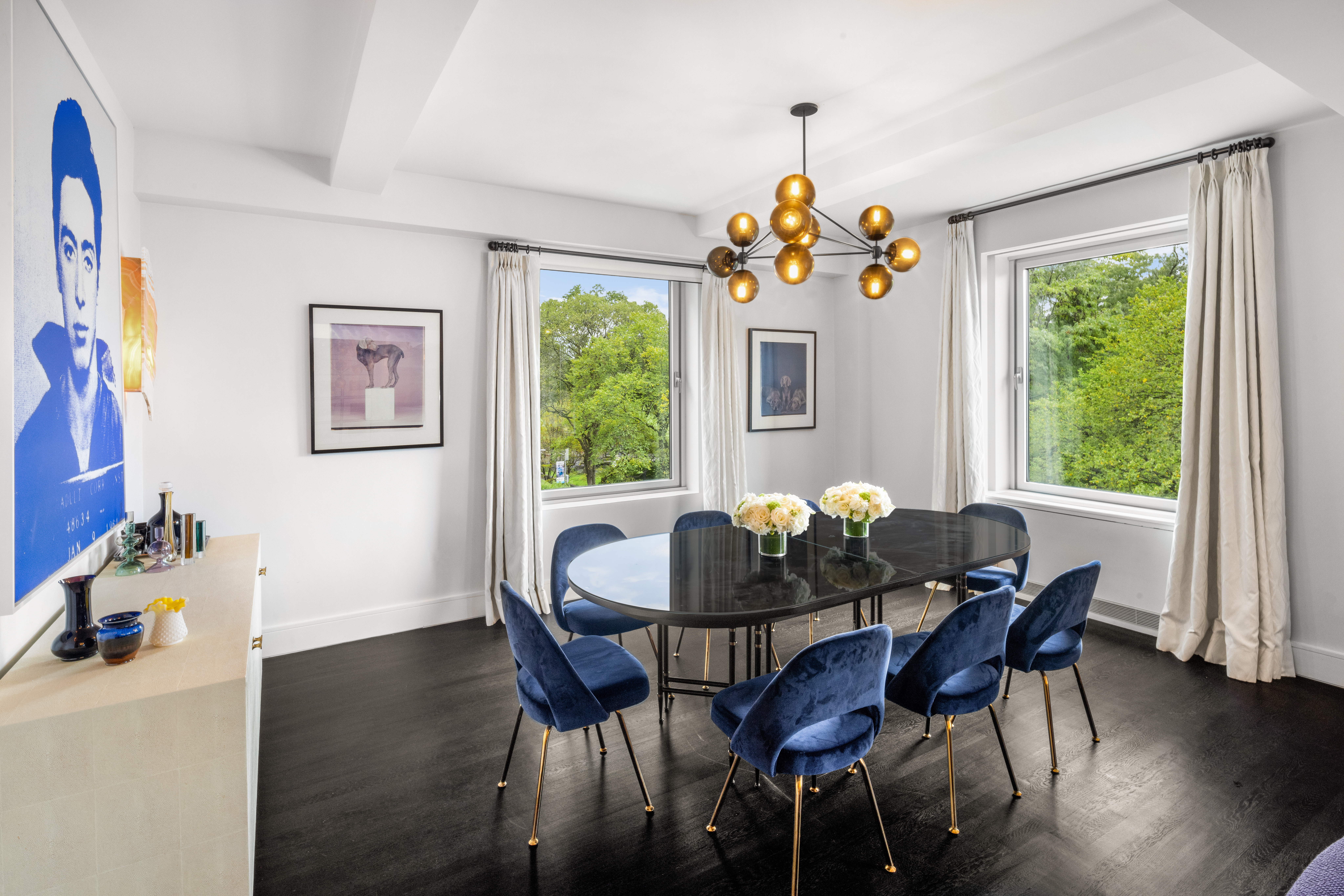 A modern dining room with a dark wooden floor, a black oval table surrounded by eight blue velvet chairs, two large windows with white curtains, a gold chandelier with spherical lights, and wall art including a portrait of James Dean.