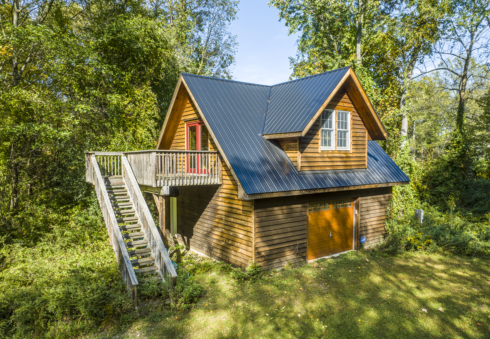 Two-story wooden cabin with a metal roof, surrounded by lush green forest and grassy ground, featuring a small upper window and a large wooden door below.