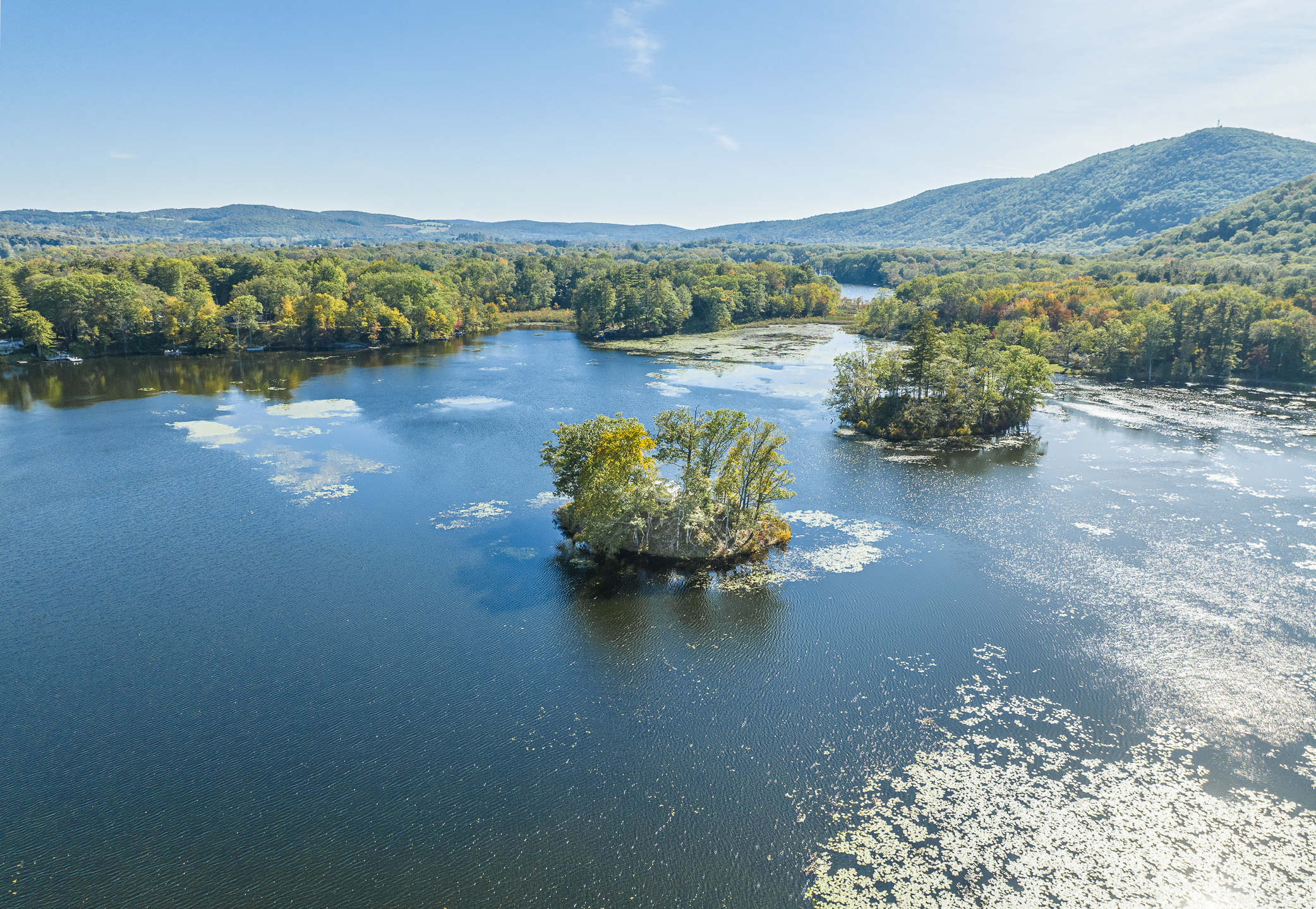 Serene lake with small tree-covered islands, surrounded by lush green forests and rolling hills under a clear blue sky.