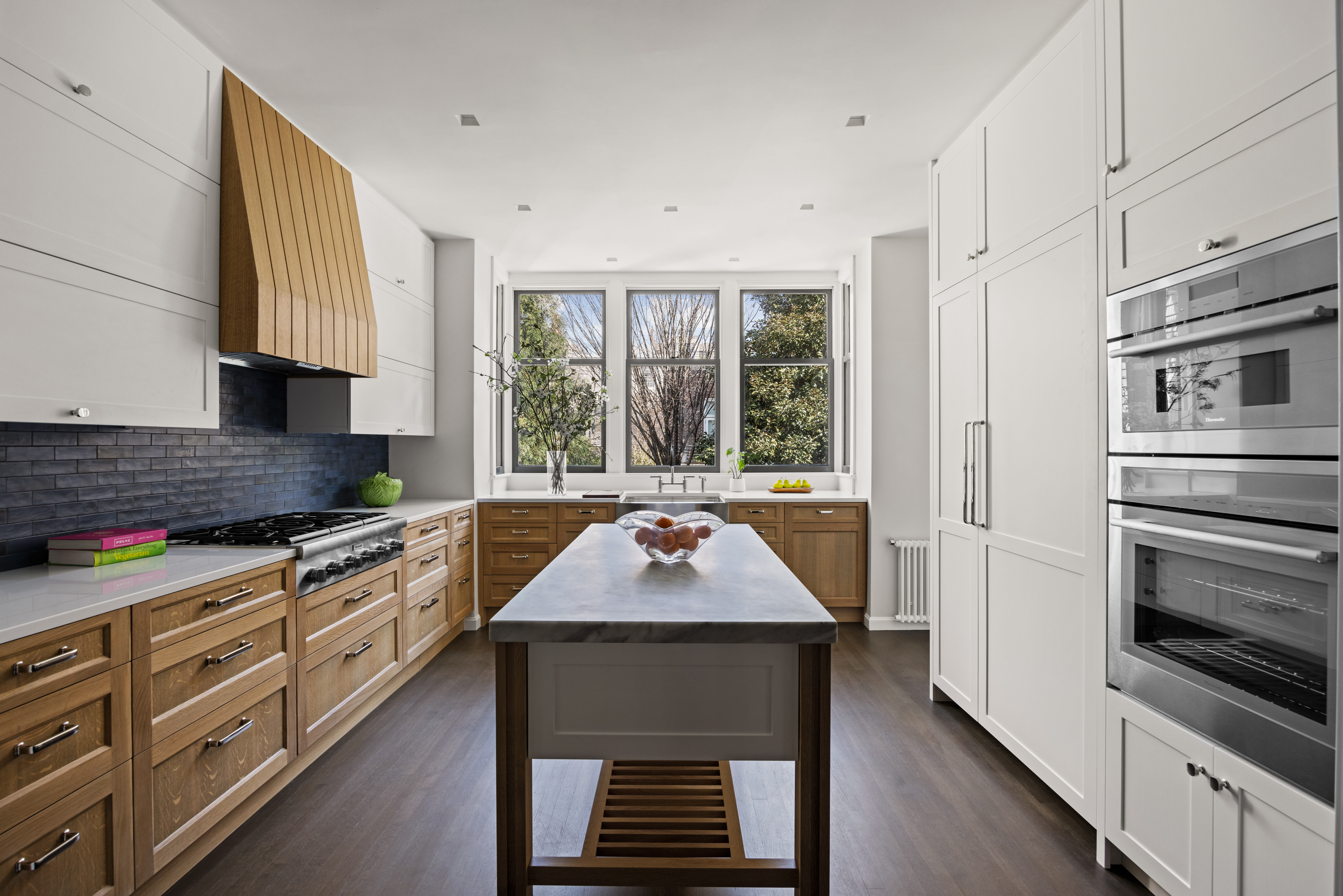 Modern kitchen with wooden and white cabinetry, a central island with a bowl of round objects, stainless steel appliances, and large windows overlooking greenery.