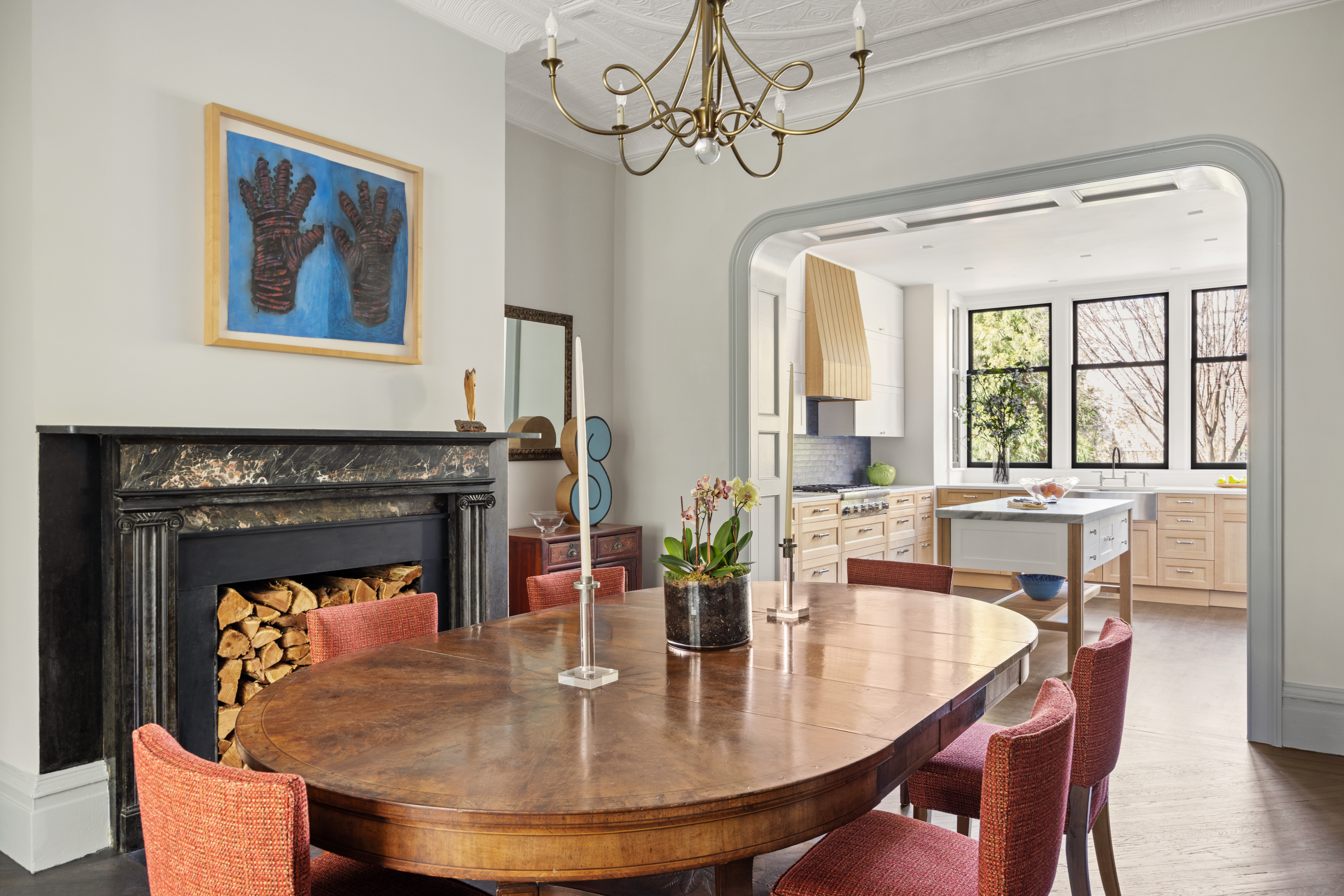 Dining room with a wooden table and red upholstered chairs, a black marble fireplace with stacked firewood and glove artwork above, and an adjacent modern kitchen with light wood cabinetry and large windows.