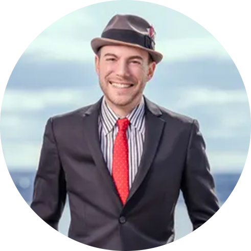 A person wearing a dark suit, striped shirt, red tie, and gray fedora stands outdoors with a blurred background.