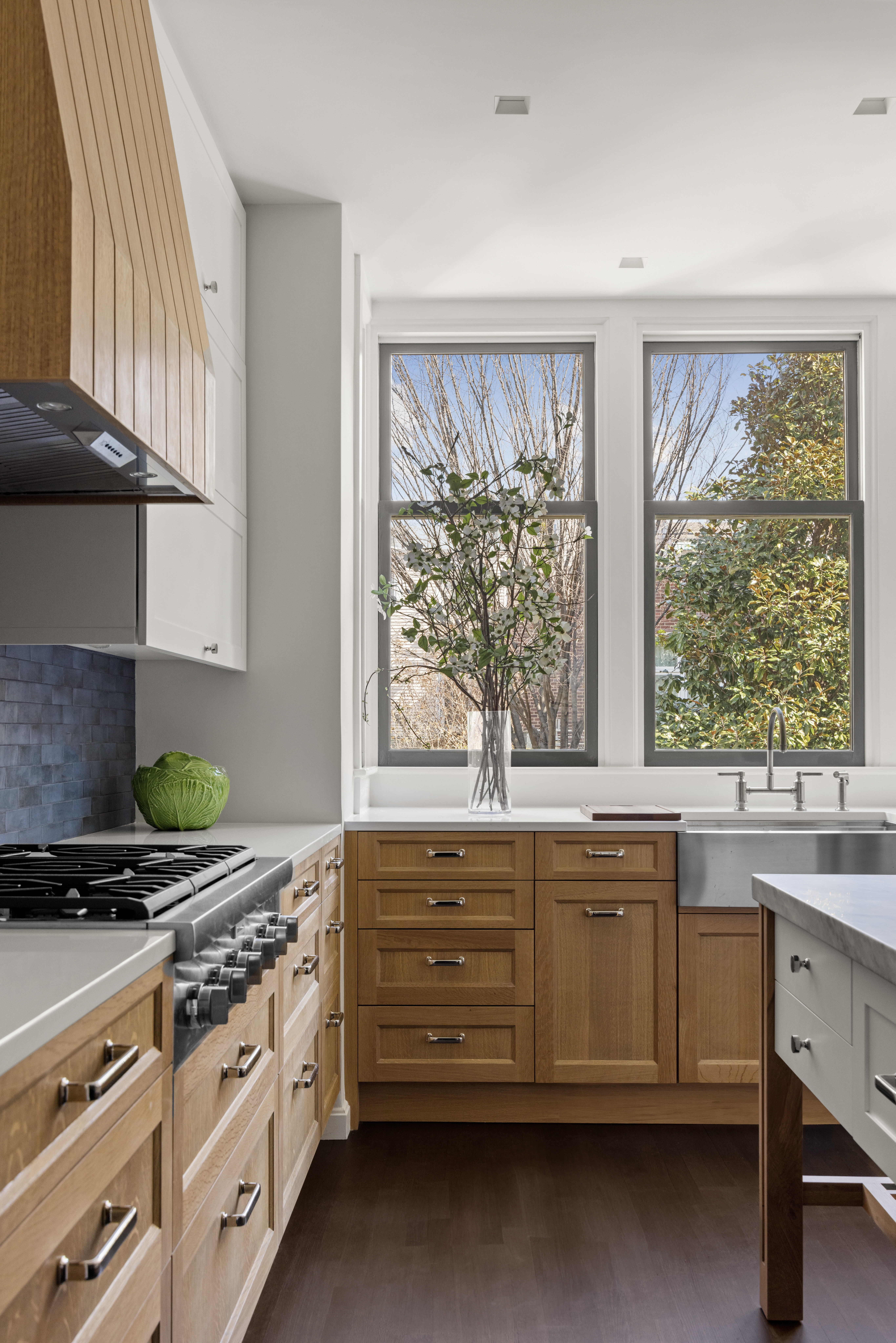 Modern kitchen with light wood cabinets, a stainless steel gas stove, and a large farmhouse sink under two windows, with a vase of branches on the countertop.