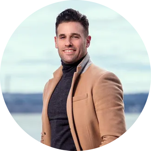 A person wearing a tan coat over a dark turtleneck sweater stands outdoors, with a blurred background of water and sky.