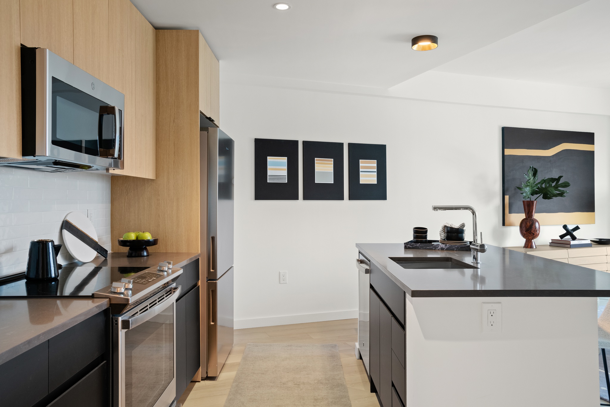 Modern kitchen with light wood cabinets, stainless steel appliances, a central island with a sink, minimalist decor, three framed abstract artworks, and a vase with green leaves on the counter.