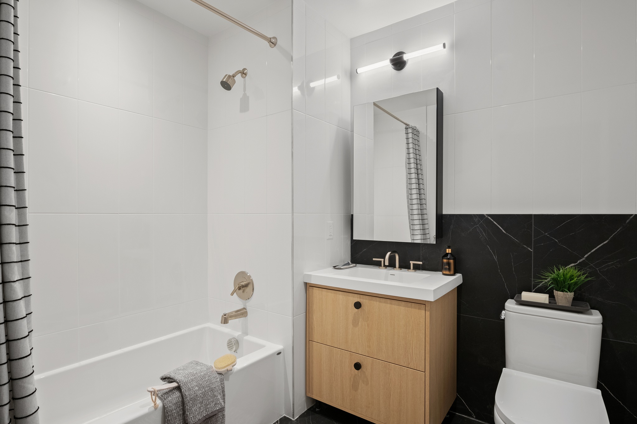 Modern bathroom featuring a white bathtub with a gray towel and beige bath brush, a wooden vanity with a white sink and rectangular mirror, and a toilet with decorative items on top.