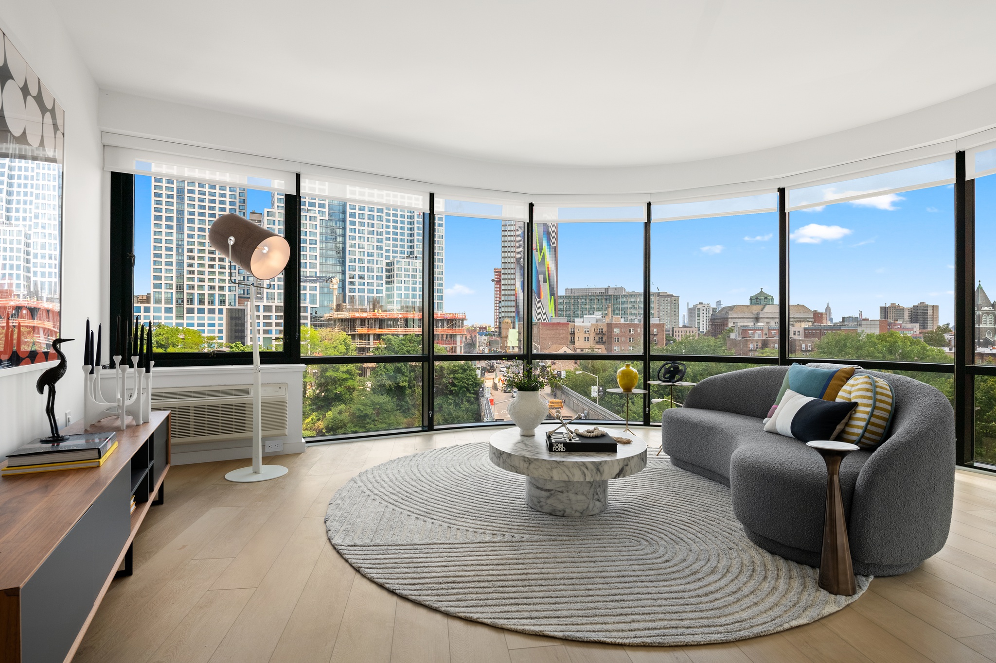 Modern living room with a curved gray sofa, colorful pillows, a round marble coffee table on a textured circular rug, and floor-to-ceiling windows overlooking city buildings and greenery