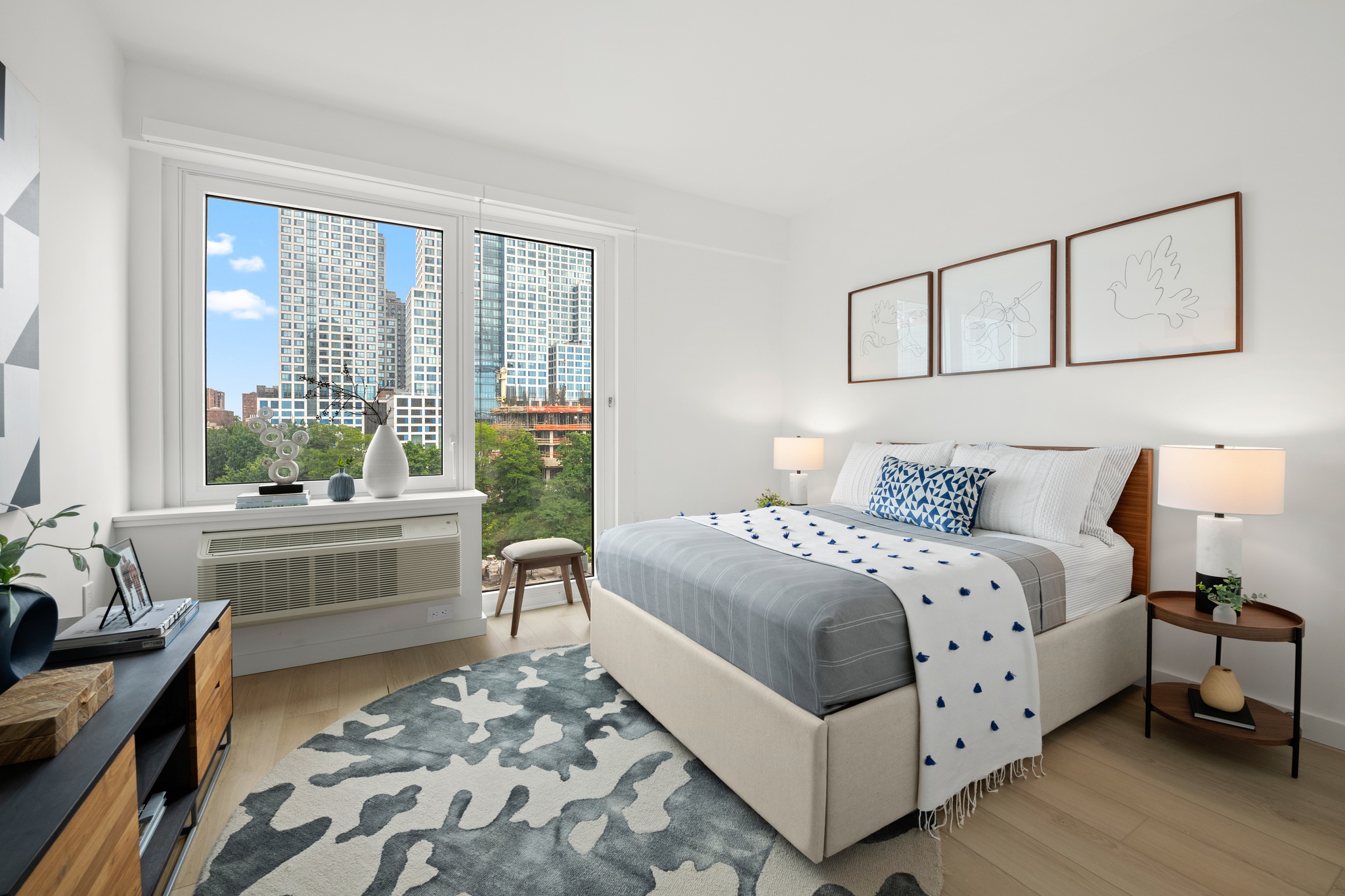 Modern bedroom with a large window showing a cityscape view, a beige upholstered bed with gray and white bedding, two bedside tables with lamps, and three minimalist bird-themed artworks above the bed.