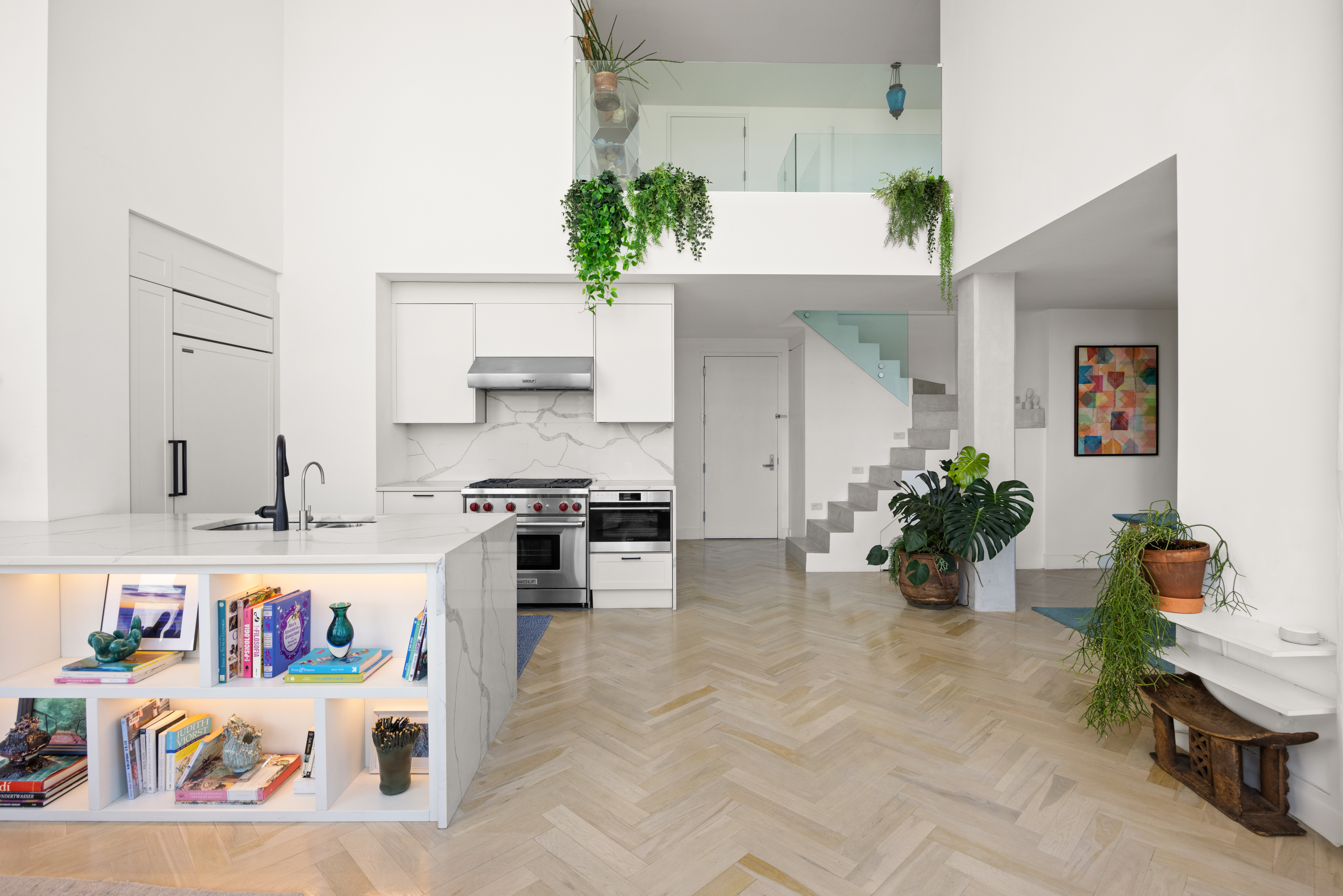 A modern, open-concept kitchen and living area with white walls and light wood herringbone floors, featuring a large island with built-in shelves, stainless steel appliances, and a marble backsplash; the living space includes plants, a staircase with a glass railing, and a door in the background.