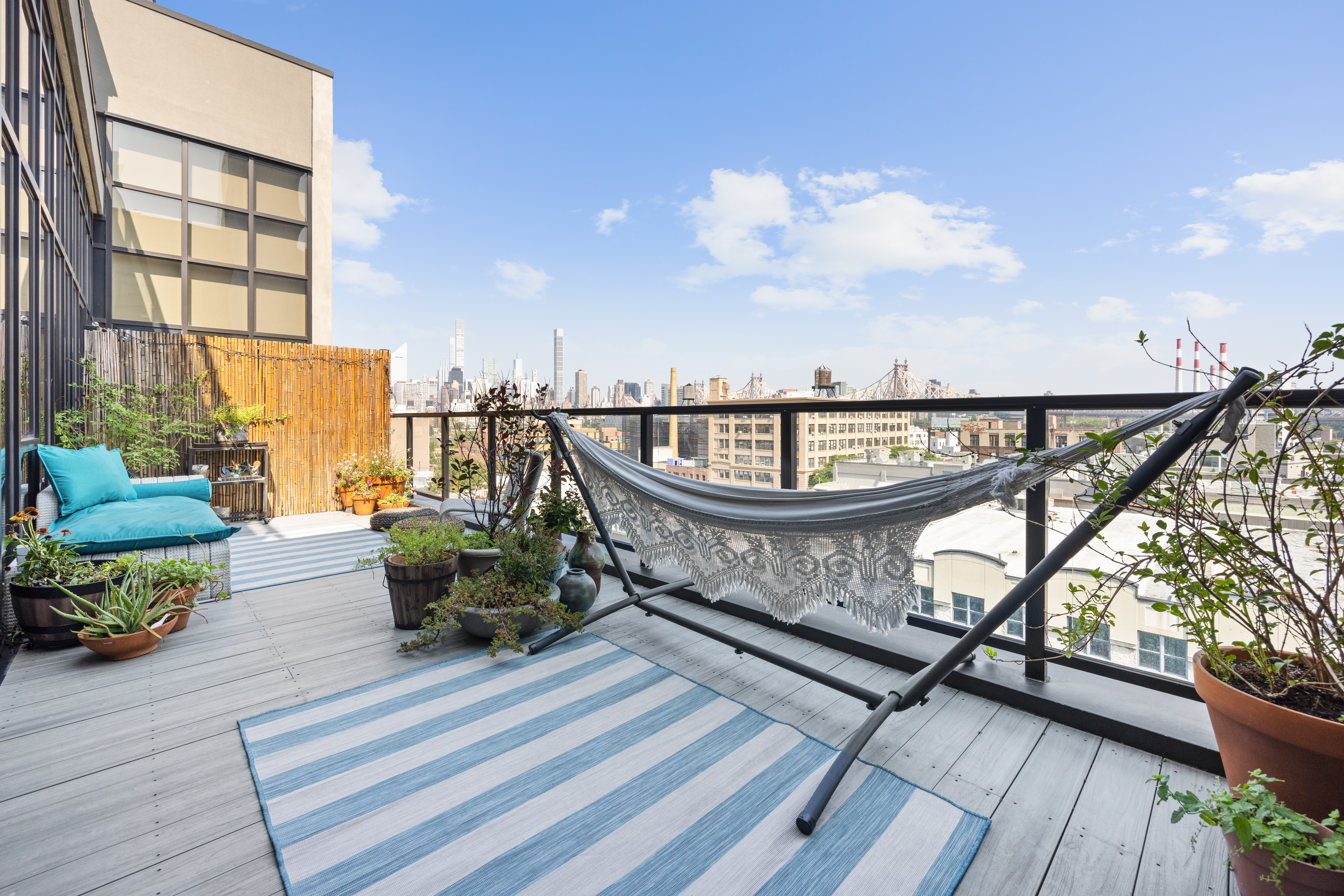 A rooftop balcony featuring a hammock, potted plants, and outdoor seating, with a view of a cityscape beneath a clear blue sky.