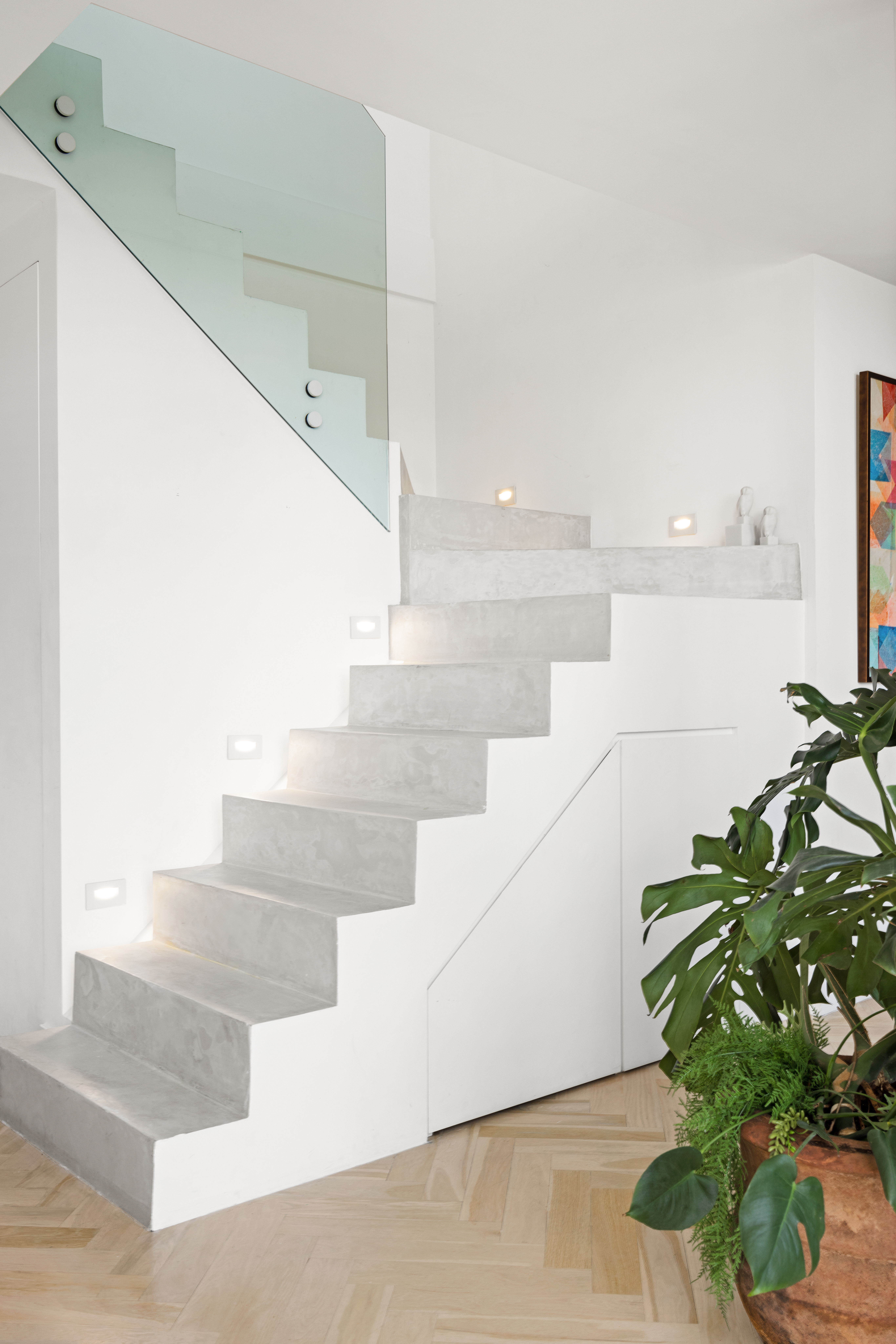 A modern, minimalist staircase with light gray steps and a glass railing, lit by small built-in side lights, with a large green plant at the base adding contrast to the clean white interior.