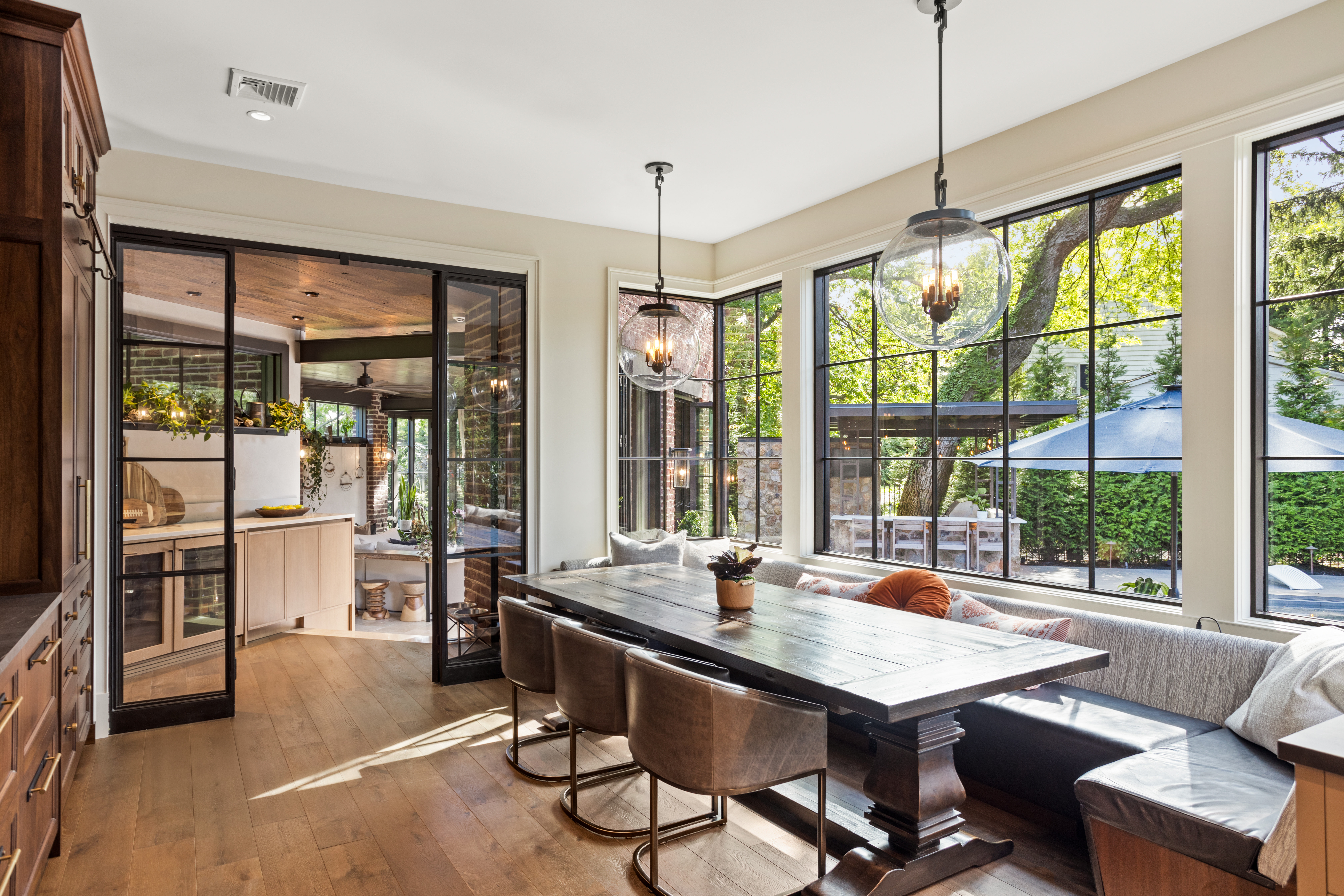 A modern dining area with a large wooden table, leather chairs, and a cushioned bench. Large windows fill the space with natural light and provide views of an outdoor patio with greenery and a blue umbrella.
