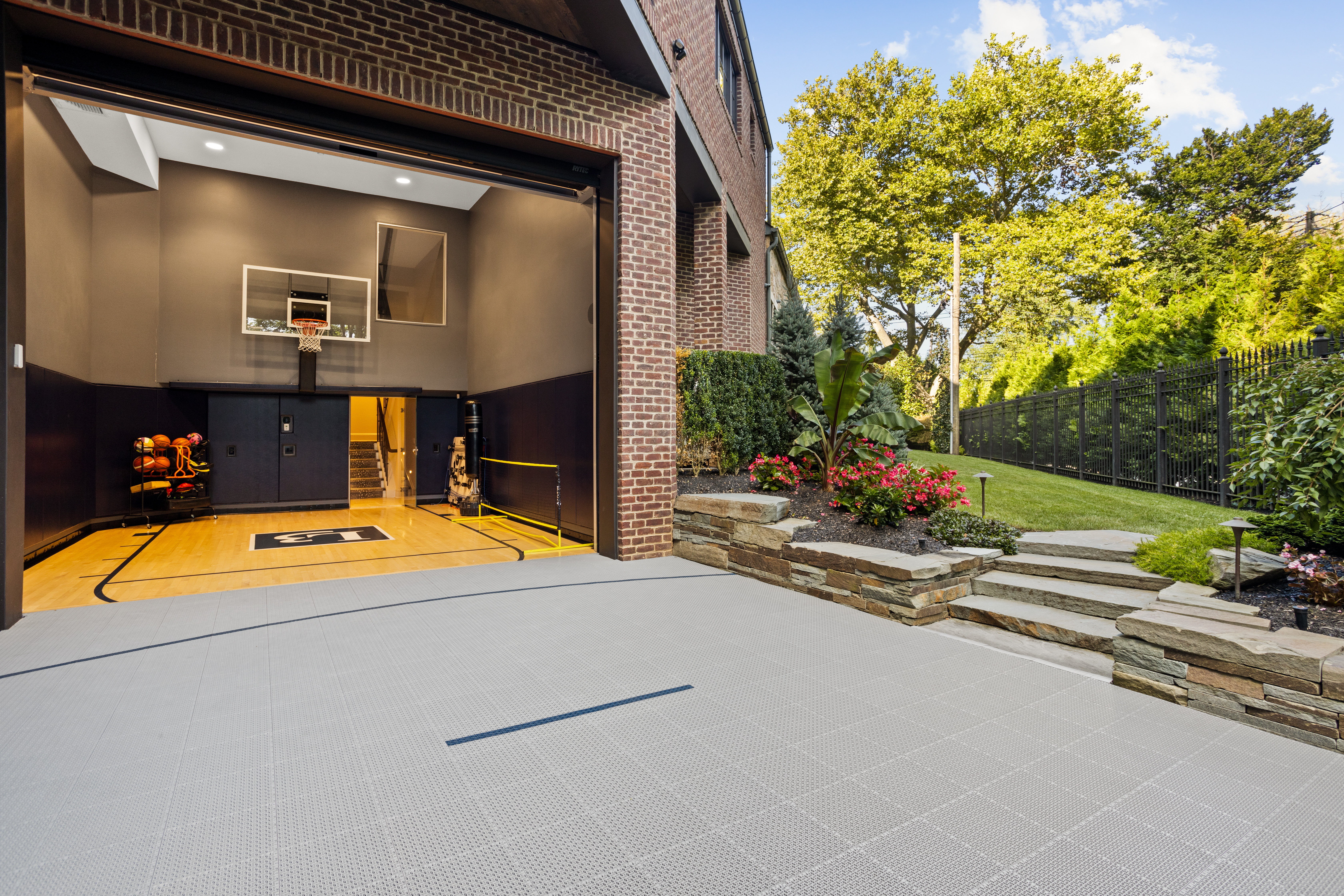 A modern home with an open garage door revealing an indoor basketball court. The exterior features a landscaped garden with stone steps leading to a grassy lawn, surrounded by trees and plants.