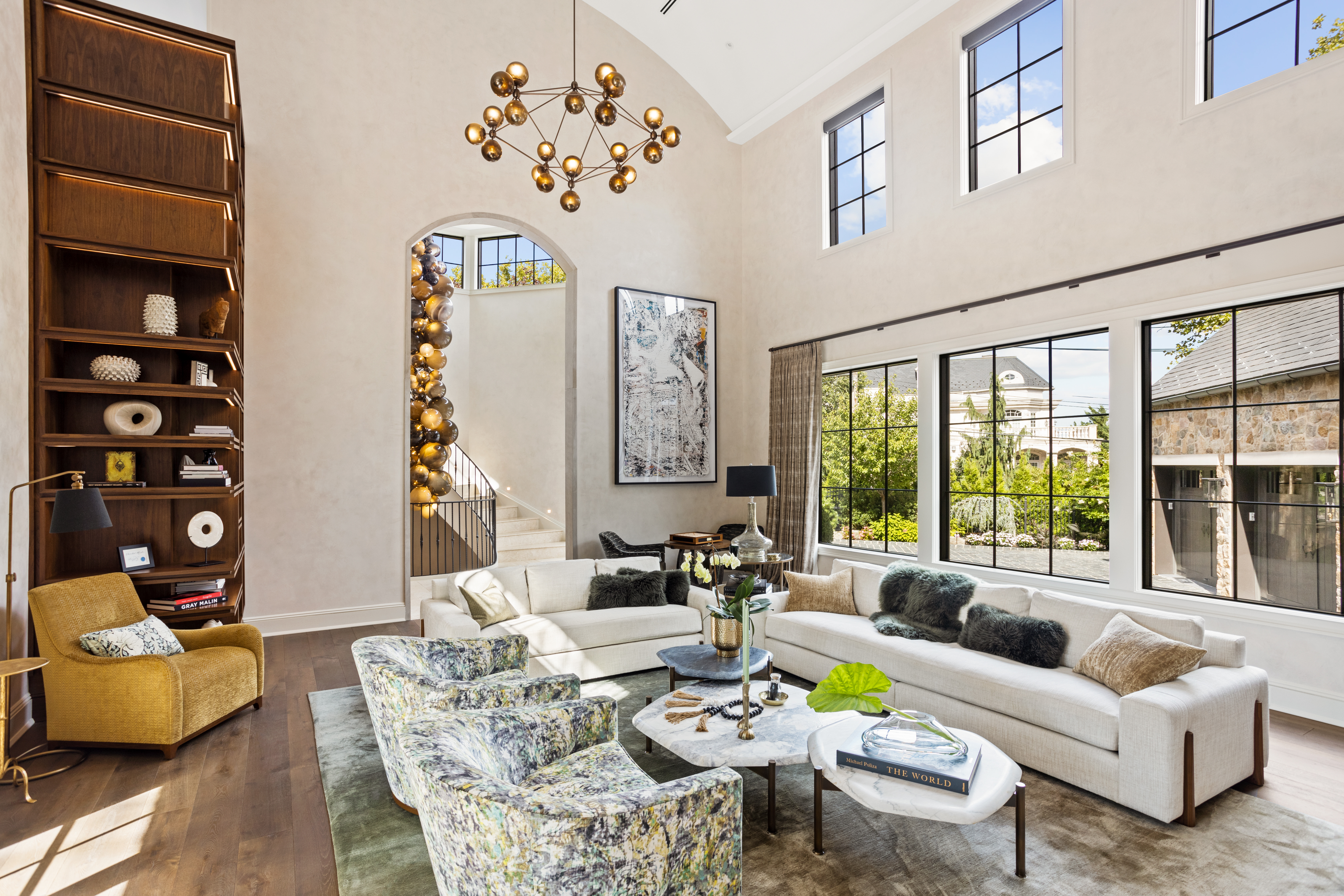 A spacious, modern living room with high ceilings, large windows, and contemporary furniture including a white sectional sofa, patterned armchairs, and a mustard yellow chair. A spherical light chandelier hangs from the ceiling, and built-in wooden shelves display decorative items.