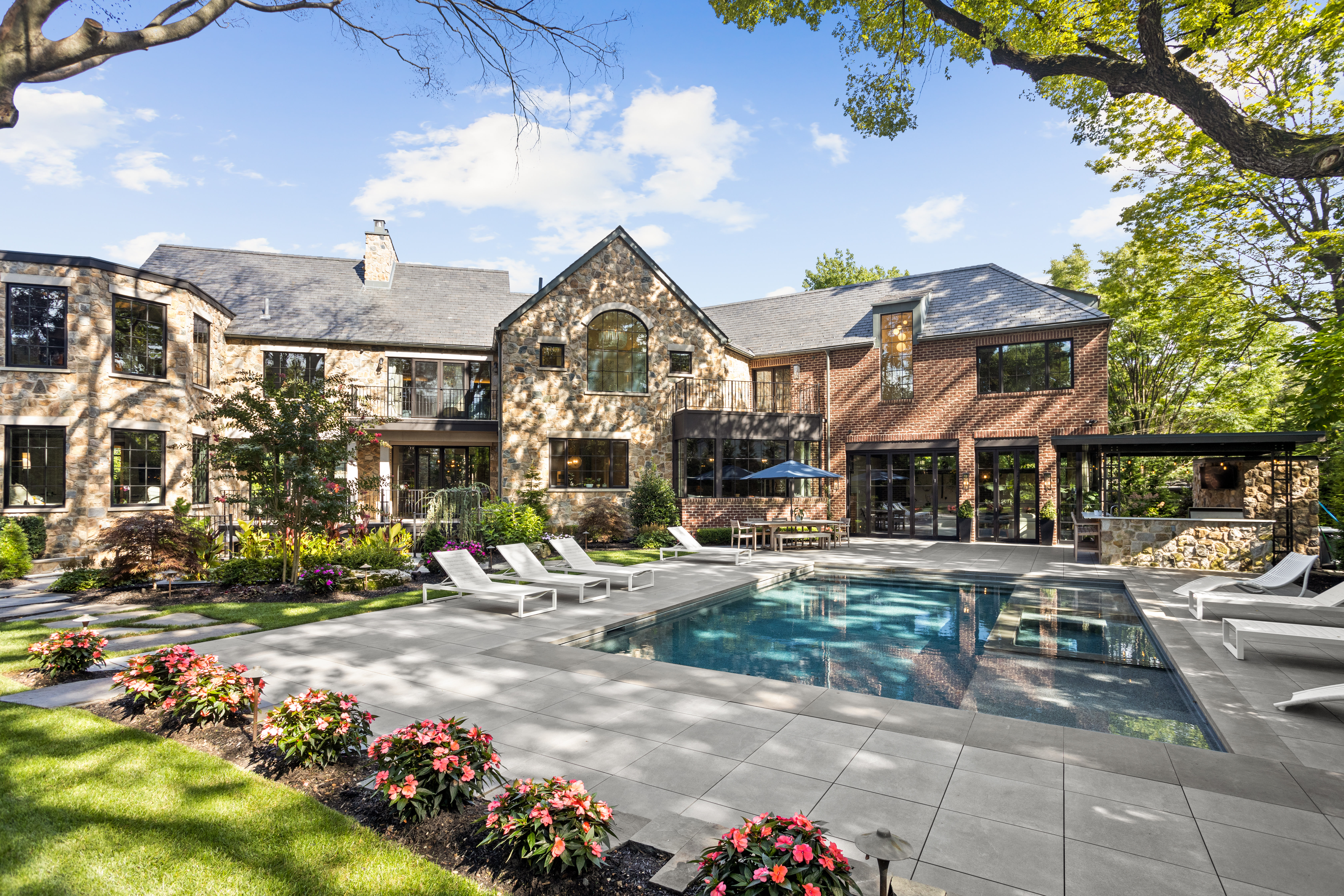 A luxurious two-story house with a stone and brick exterior, surrounded by a manicured garden. The backyard features a swimming pool with white lounge chairs, an outdoor seating area under an umbrella, and lush greenery.