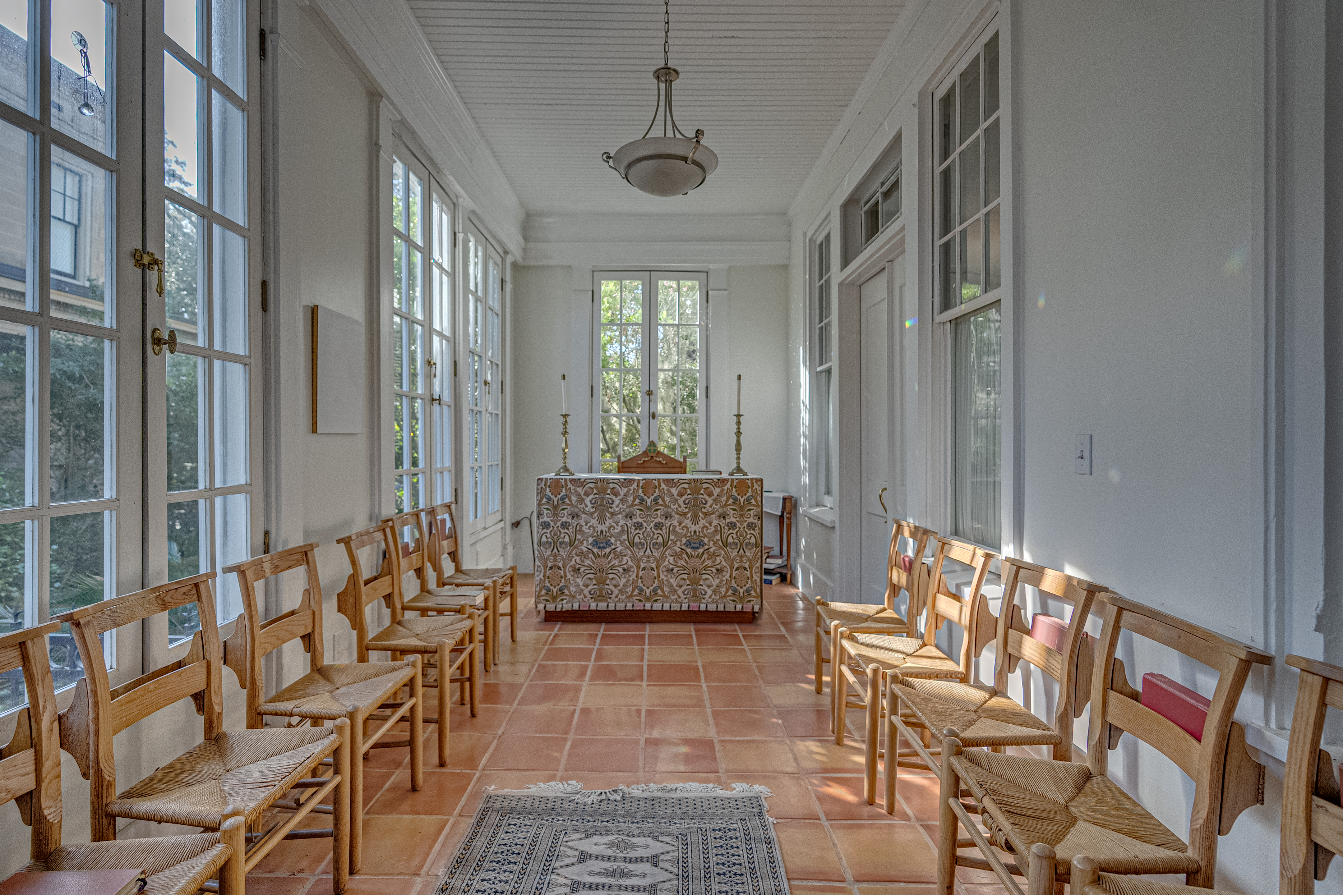 A small walkway with wooden chairs in two rows facing a tiled piece of furniture illuminated by natural light from large windows on both sides.