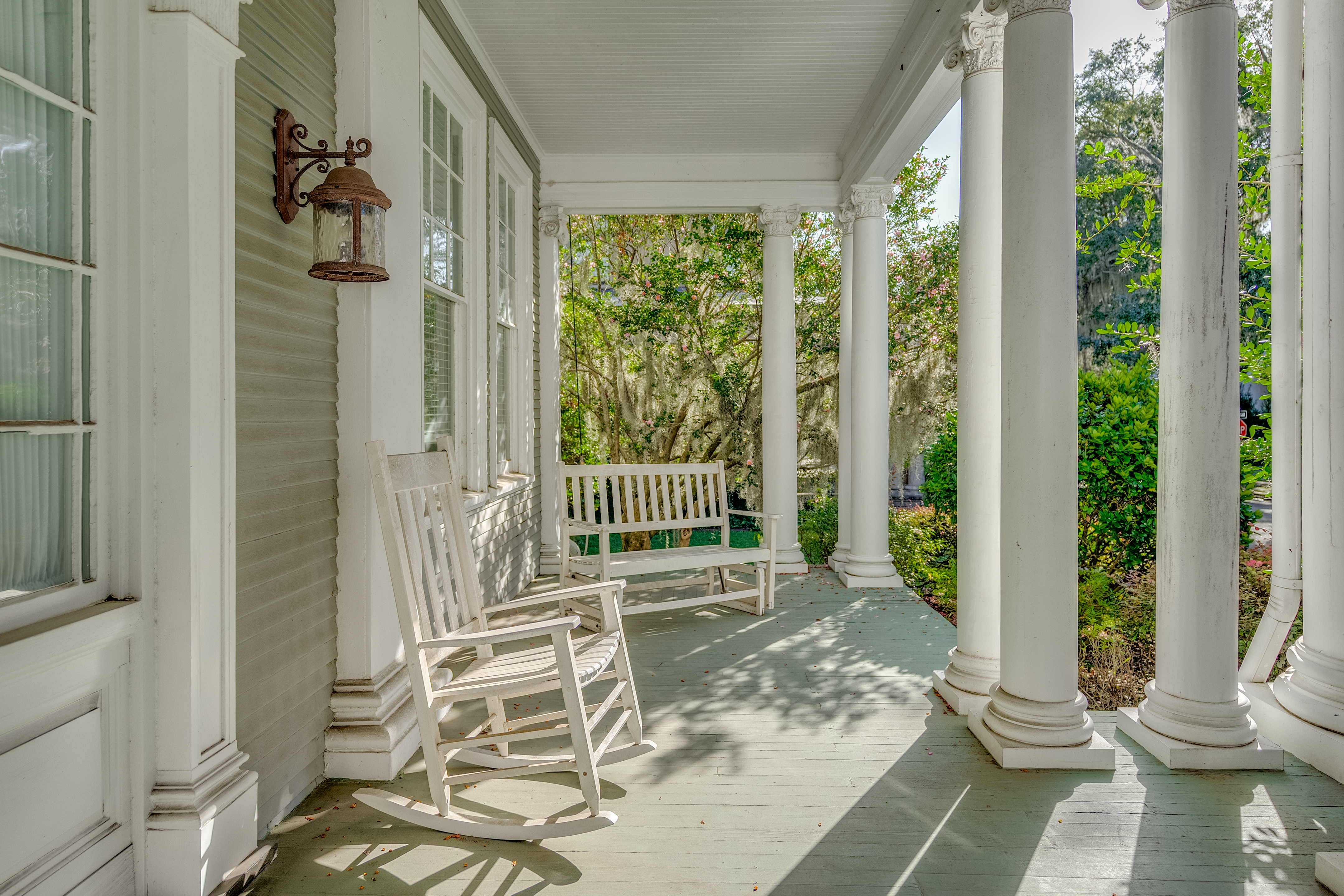 A sunlit porch with white columns, featuring a wooden rocking chair and a bench swing, overlooking a lush garden filled with trees and greenery.
