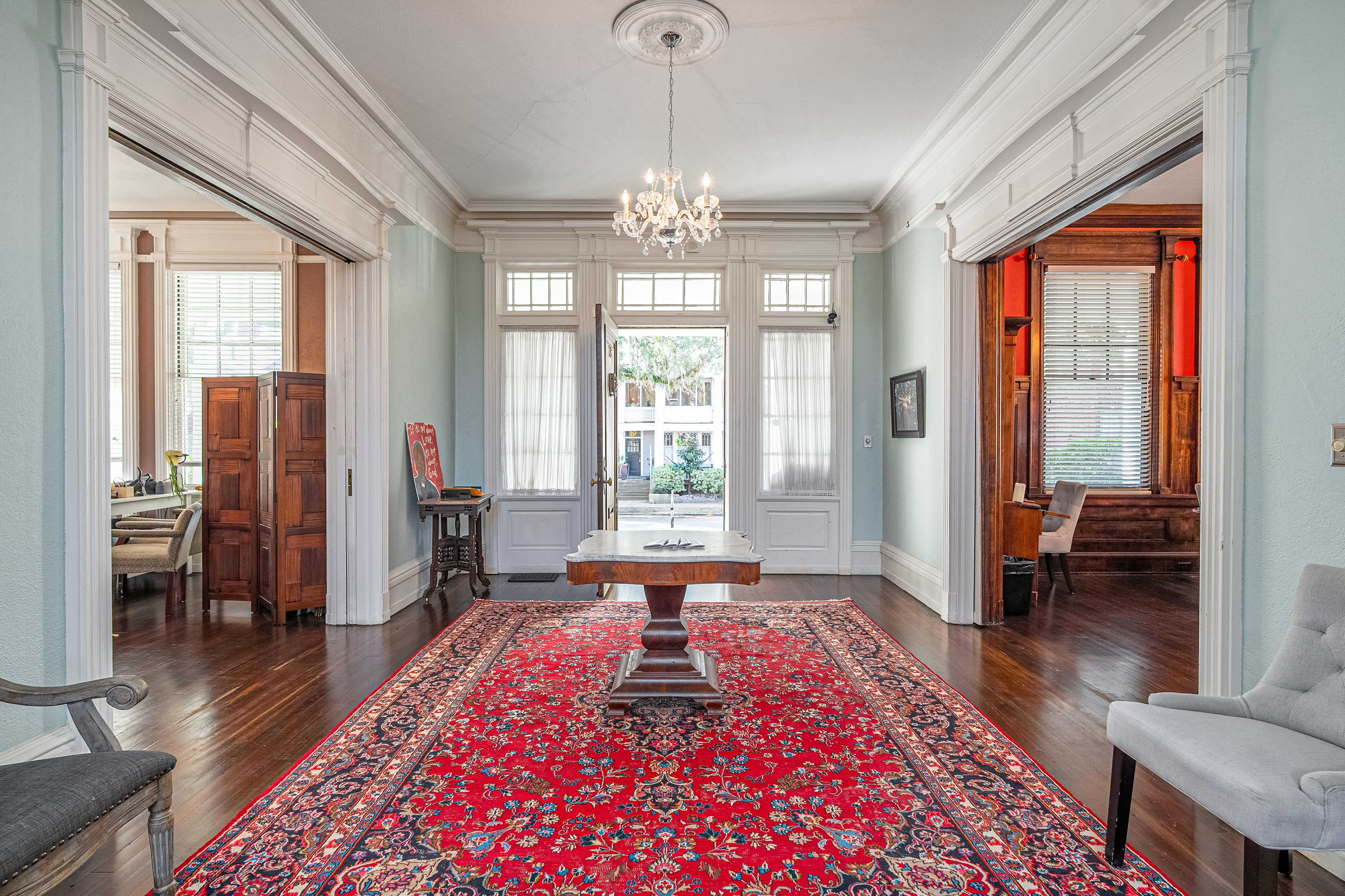 A spacious, elegant room with a large red Persian rug, a central wooden table under a chandelier, light blue walls with white trim, and multiple doorways leading to adjacent rooms.