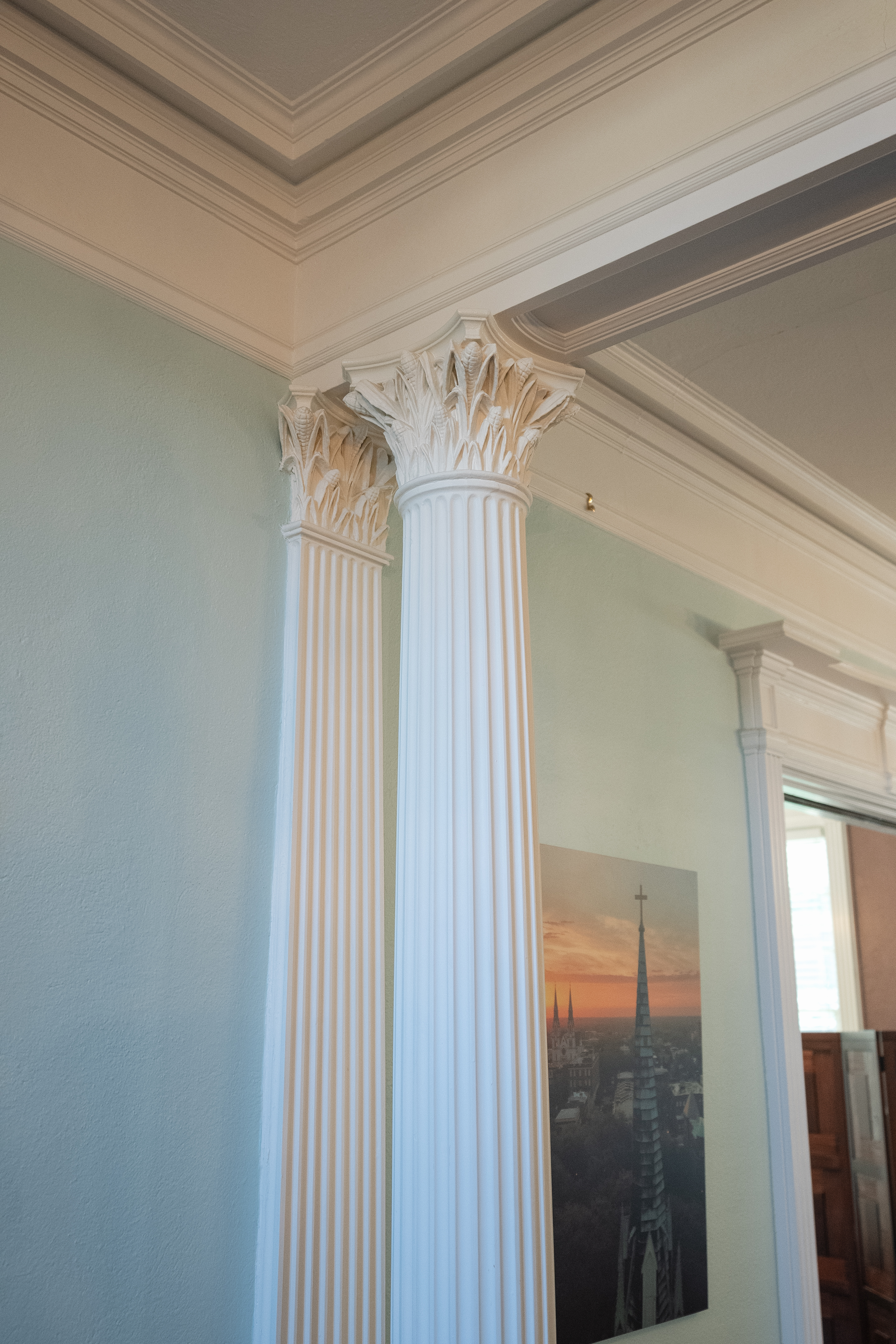 Two white Corinthian columns with ornate acanthus leaf capitals, set against a light blue wall and supporting an intricately designed ceiling.