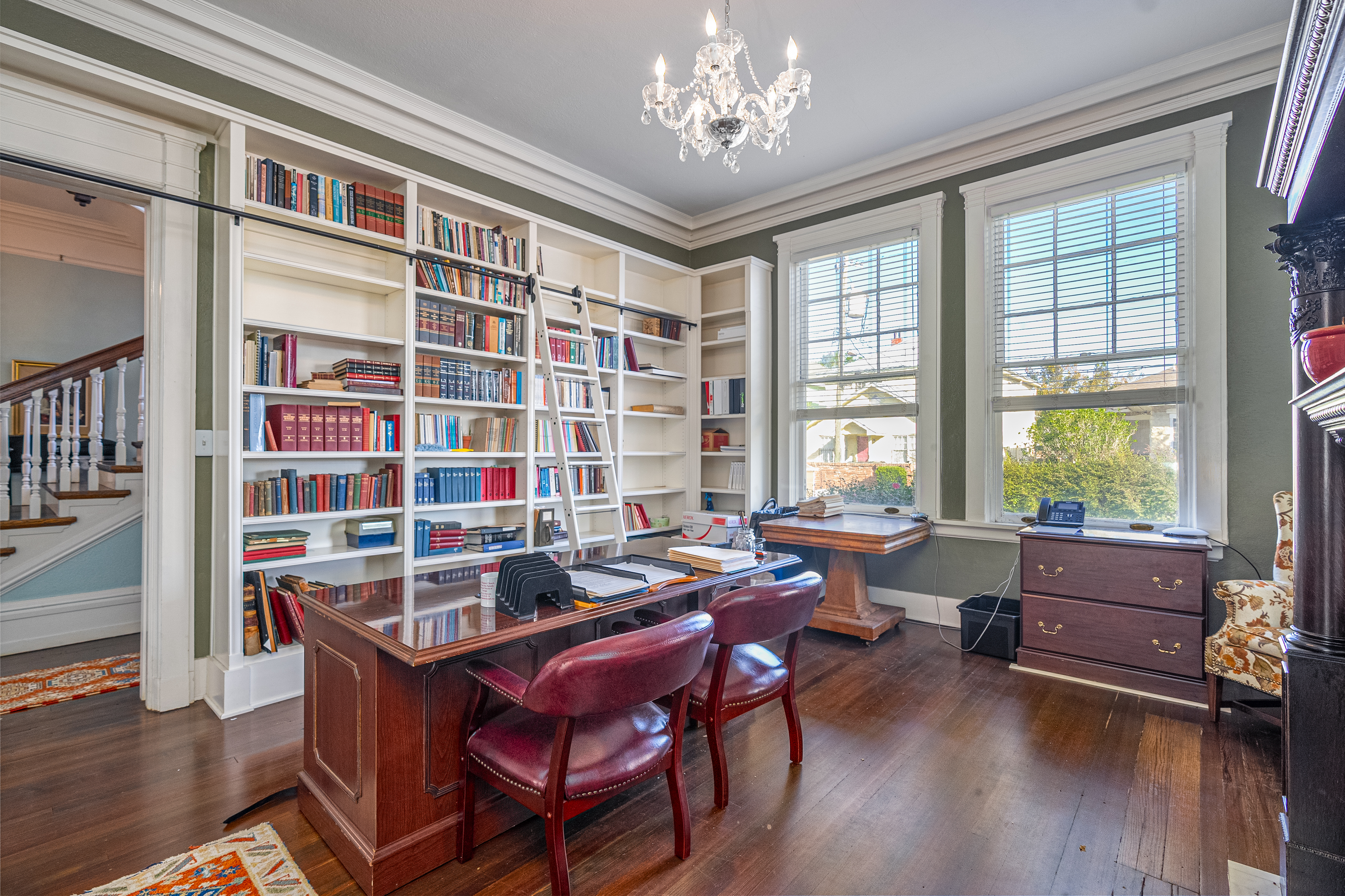 A well-lit home office with a wooden desk, two red chairs, a large bookshelf filled with books, three tall windows, a chandelier, and a ladder for reaching upper shelves.