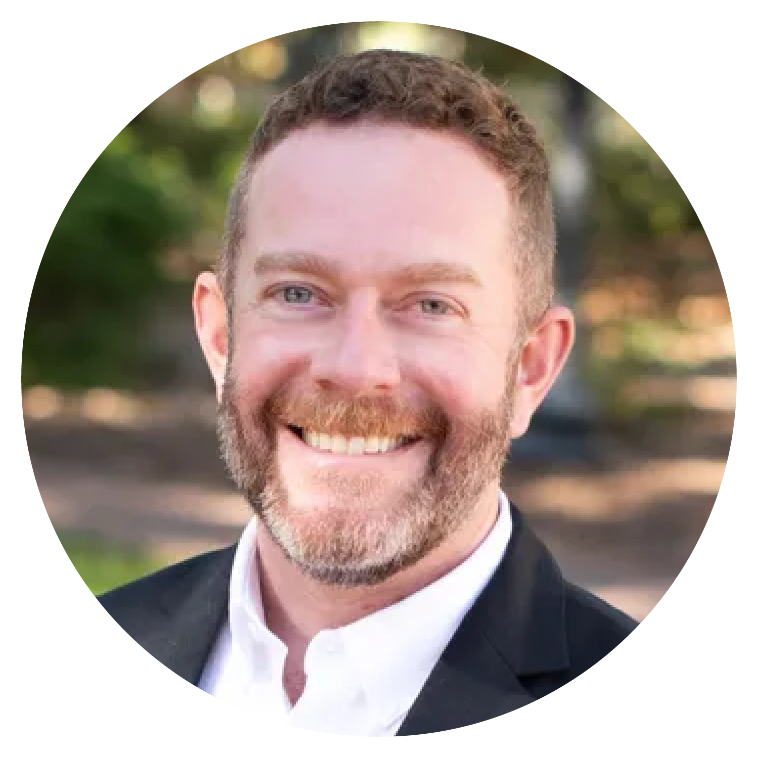 A person with short hair wearing a dark suit jacket and white shirt, standing outdoors with blurred greenery in the background.