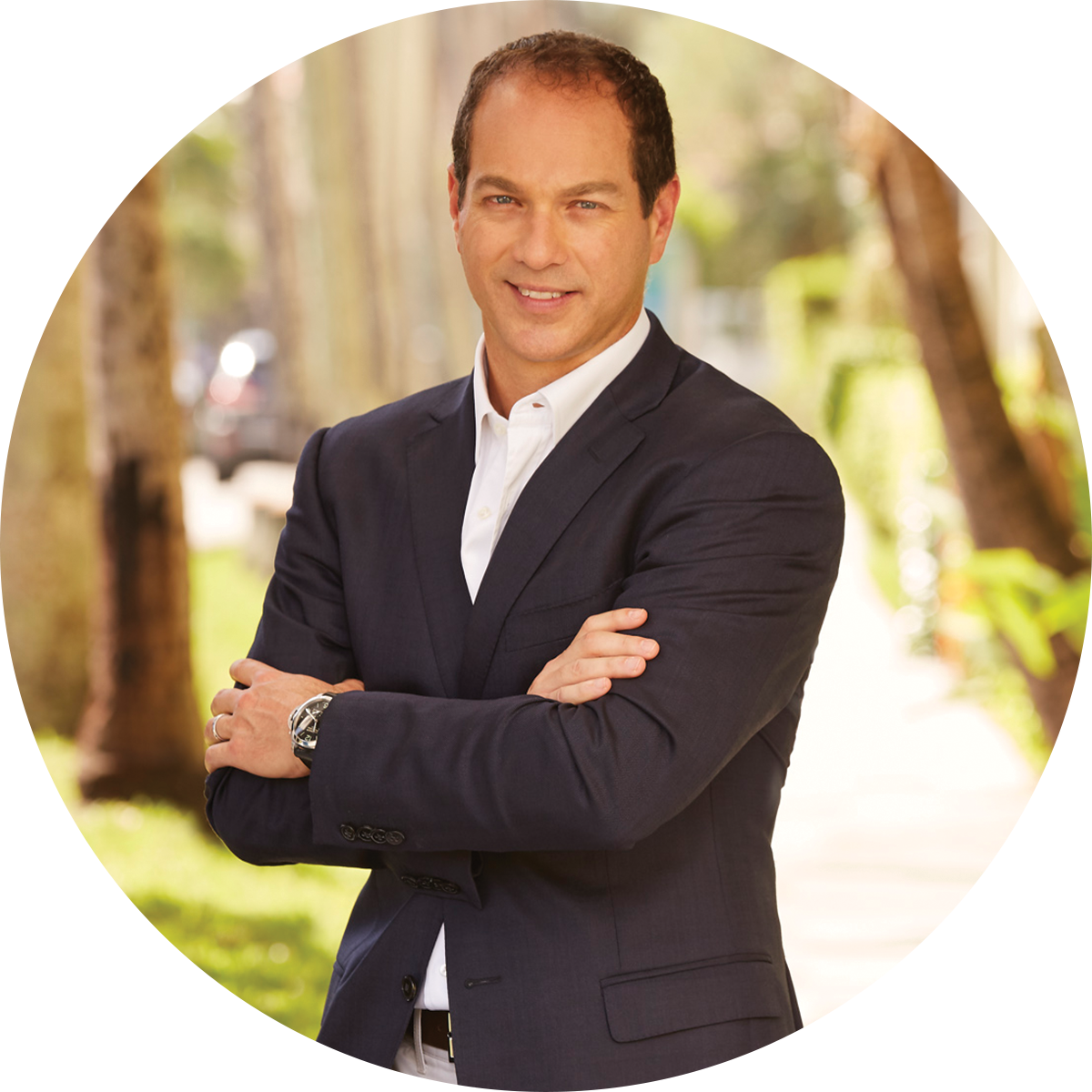 A person wearing a dark suit jacket and white shirt stands outdoors with arms crossed; the background features blurred trees, greenery, and a pathway.