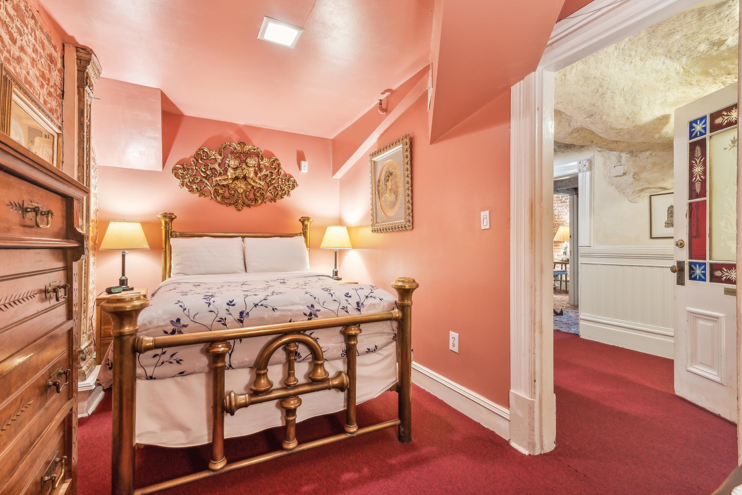 A cozy bedroom with coral-colored walls and red carpet. The room features a brass bed frame with white bedding adorned with blue floral patterns. Two bedside tables each hold a lamp with beige lampshades. Above the bed, there is an ornate gold decorative piece on the wall. To the right, an open door leads to another room with cream-colored wainscoting and a textured ceiling.