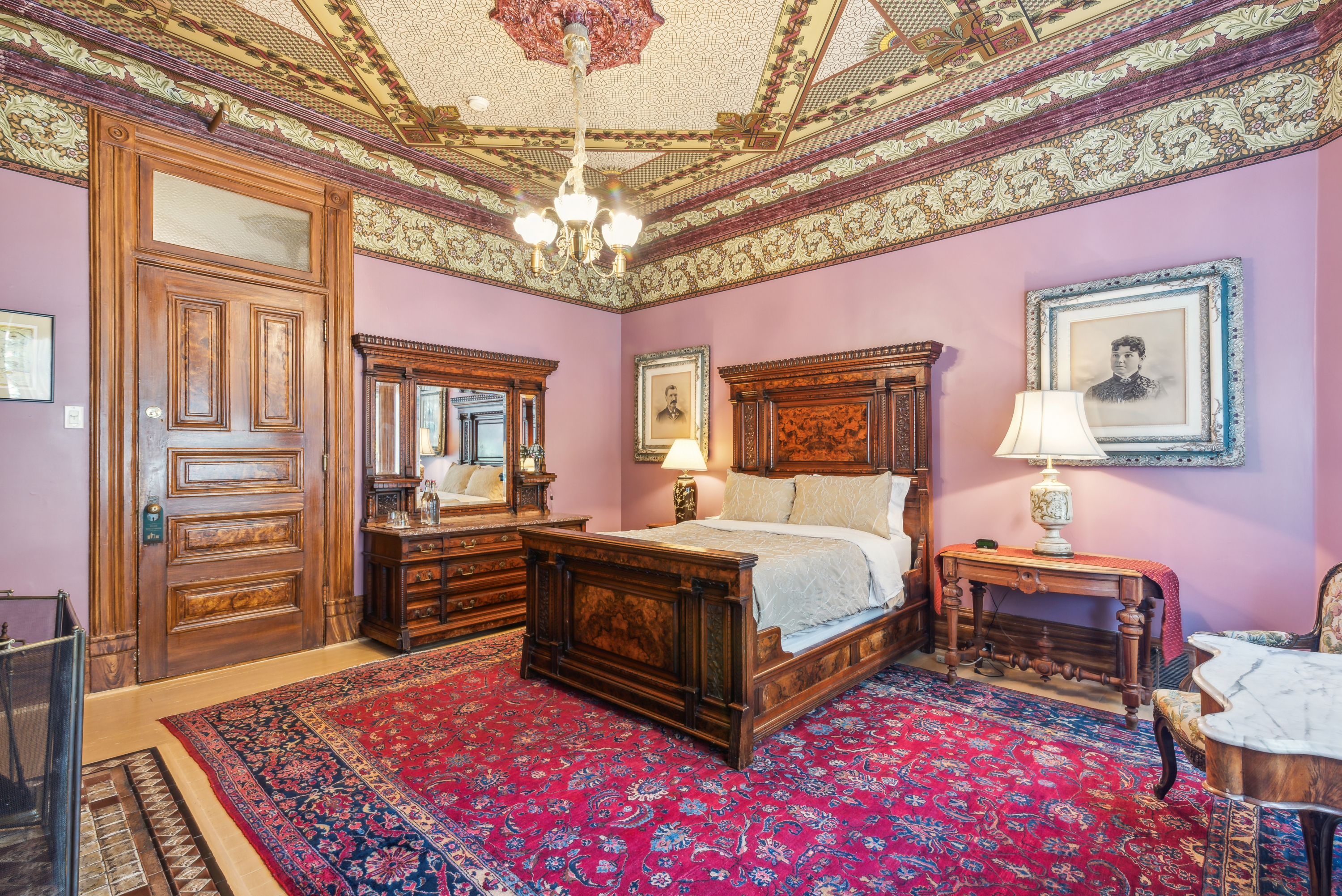 Ornate pink bedroom with decorated ceiling, hanging chandelier, and red Oriental-style rug.
