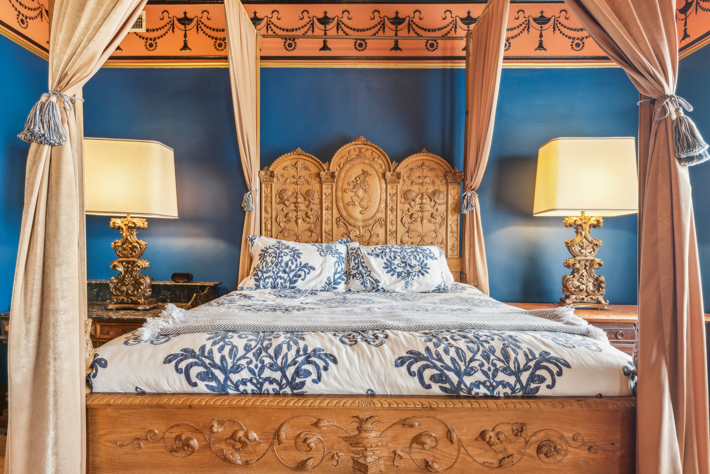 Four-poster bed with drapes and blue walls, flanked by ornate lamps on side tables.