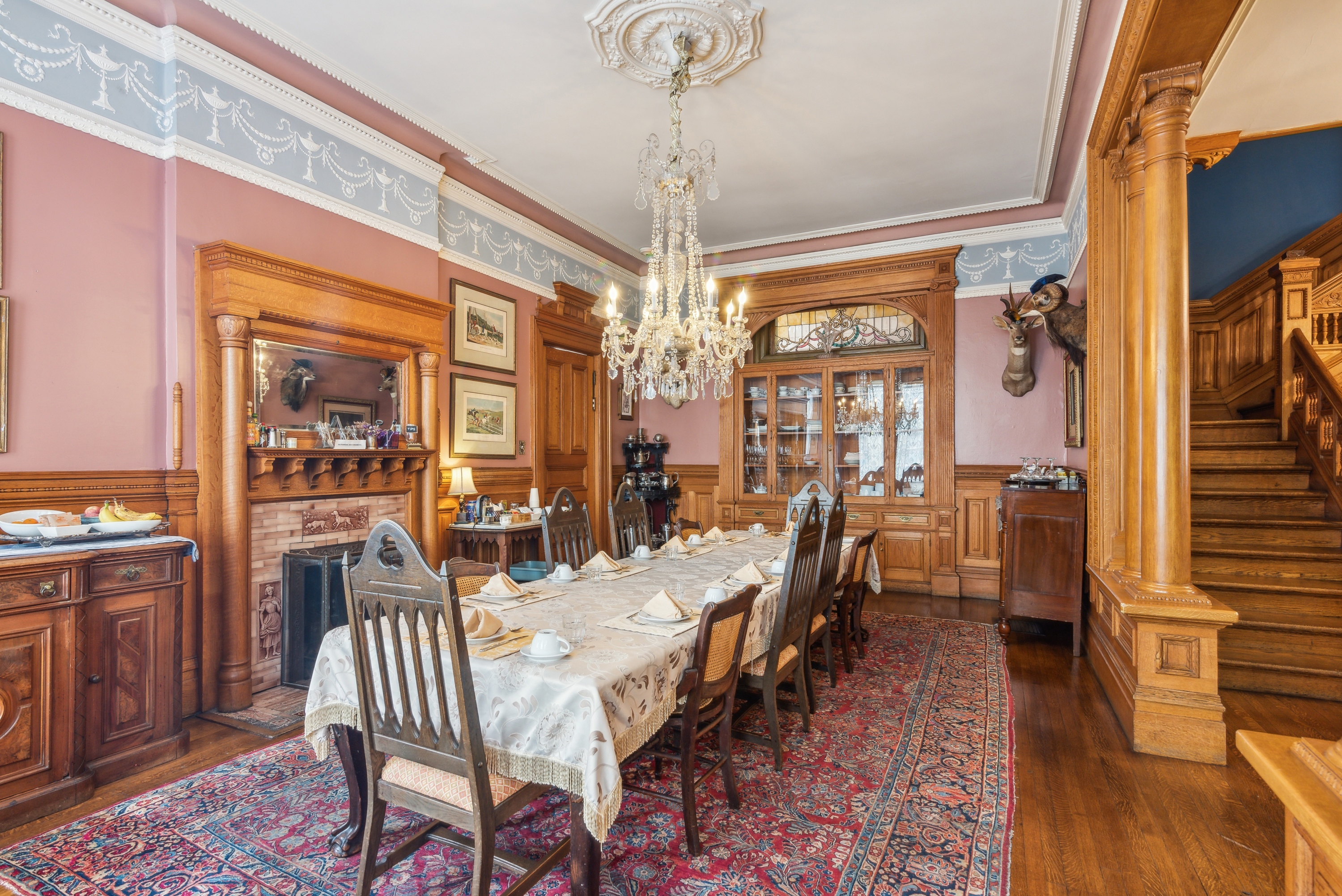 A spacious dining room with a long table covered in a white tablecloth, surrounded by wooden chairs. A chandelier hangs from the ceiling, and there are wooden paneling and decorative elements on the walls.