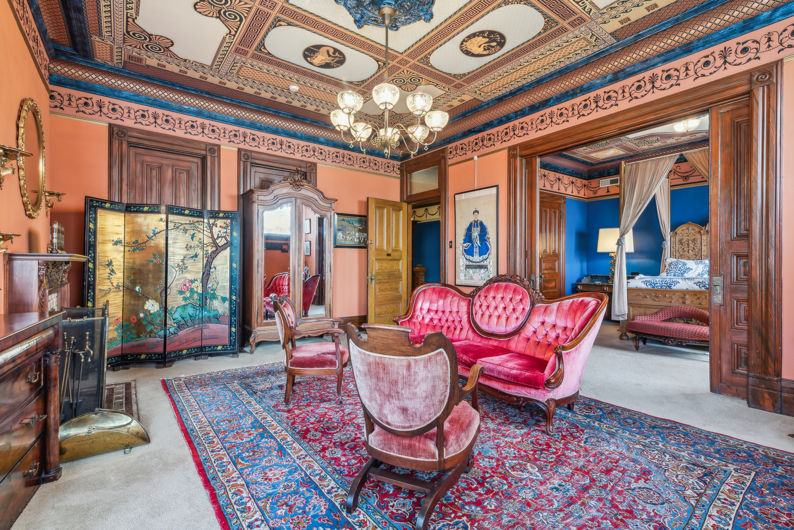 A lavishly decorated living room with ornate wooden paneling, a large chandelier, and antique furniture including red upholstered chairs and a sofa. The room features an intricate ceiling design and a colorful Persian rug on the floor.