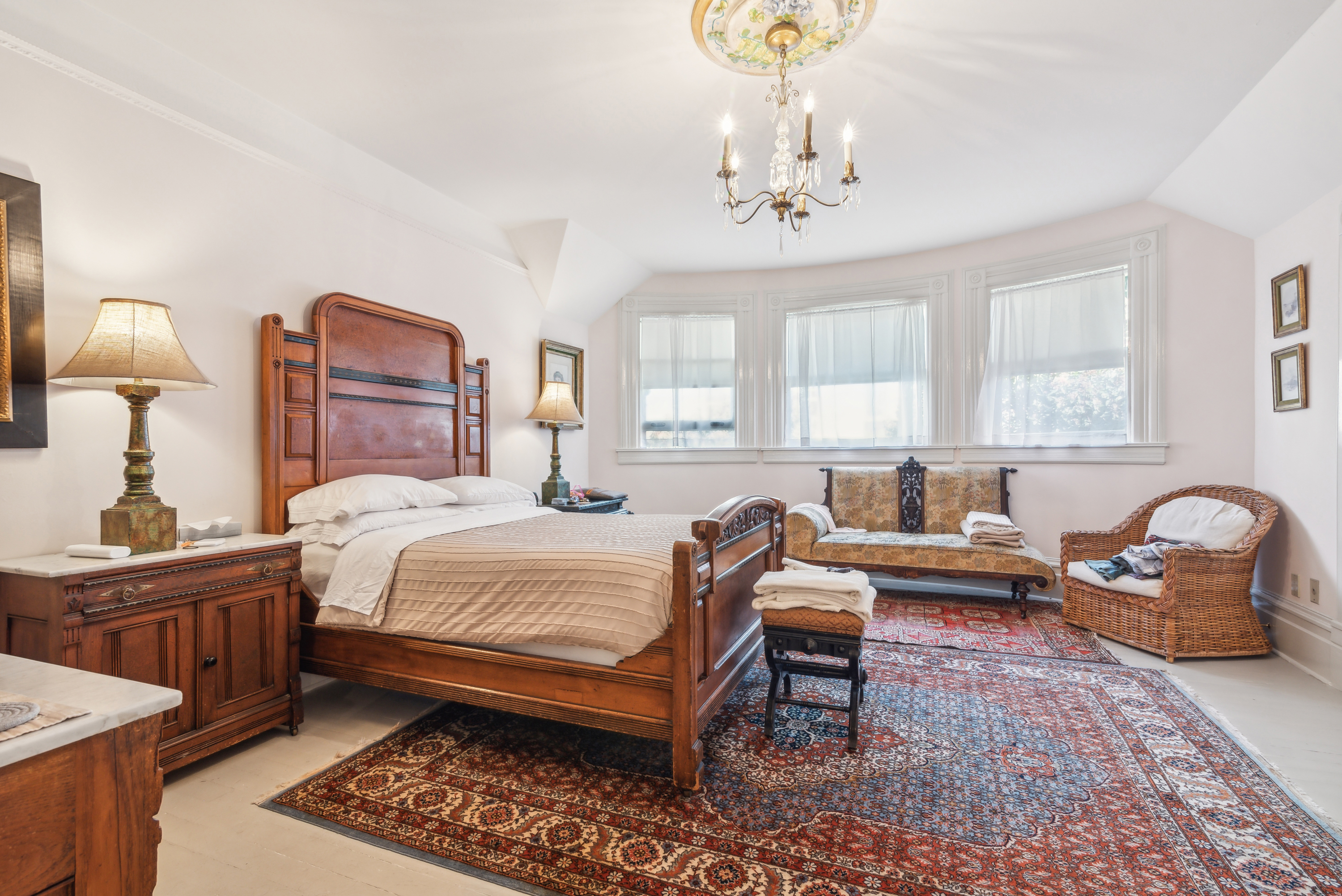 White bedroom with chandelier overhead and wooden furniture, including a bed and chairs.