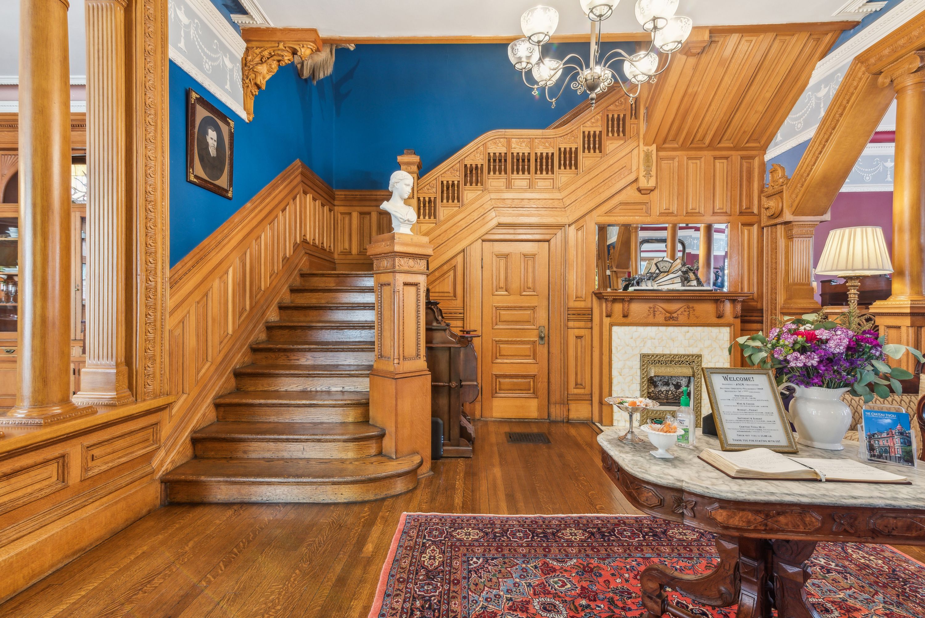 A grand staircase made of wood with ornate railings leading up to another floor. At the base of the stairs is a small table adorned with flowers.