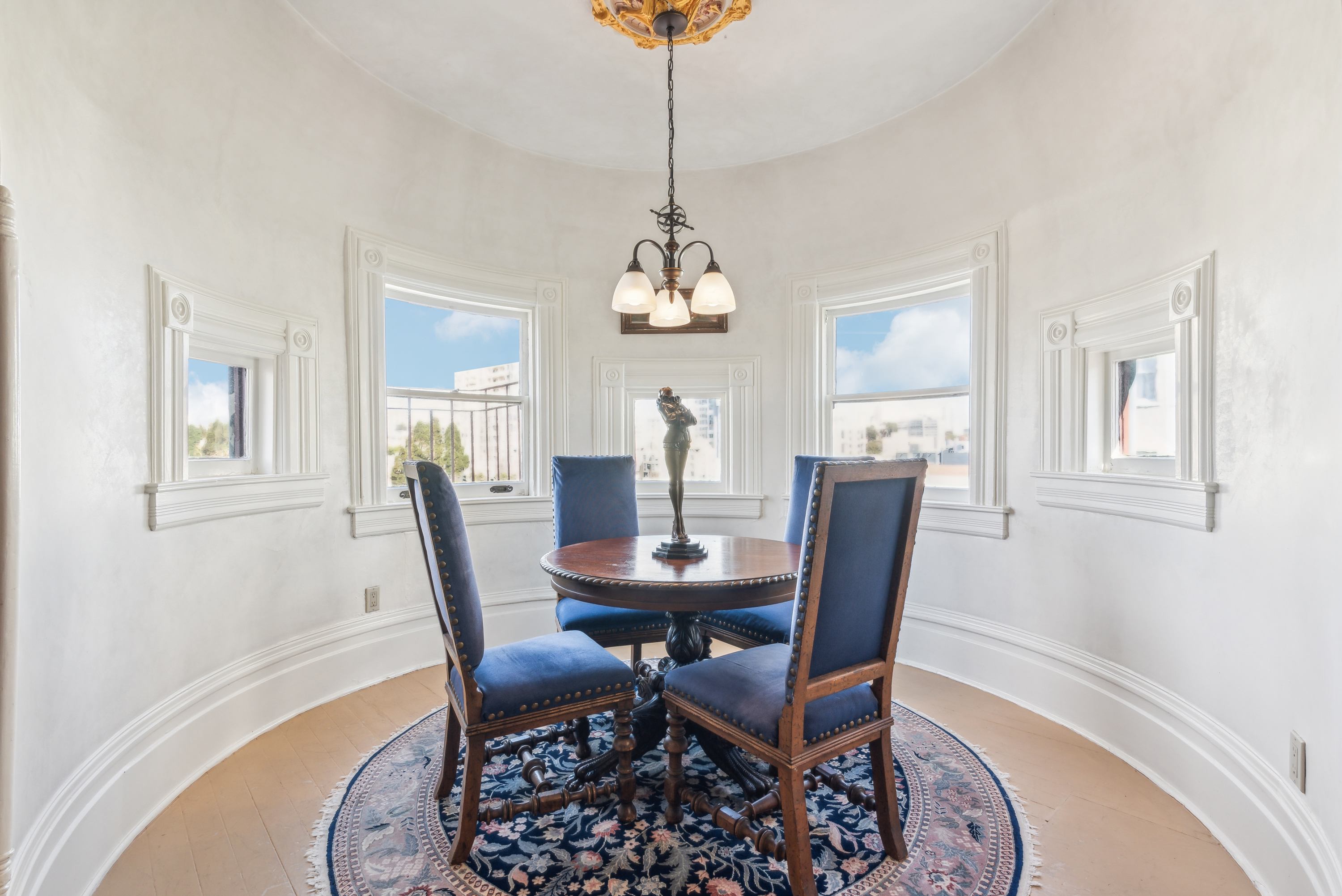 A bright dining area with white walls, featuring a round table surrounded by four dark blue upholstered chairs. The room has three small windows providing natural light and is illuminated by a hanging light fixture above the table.