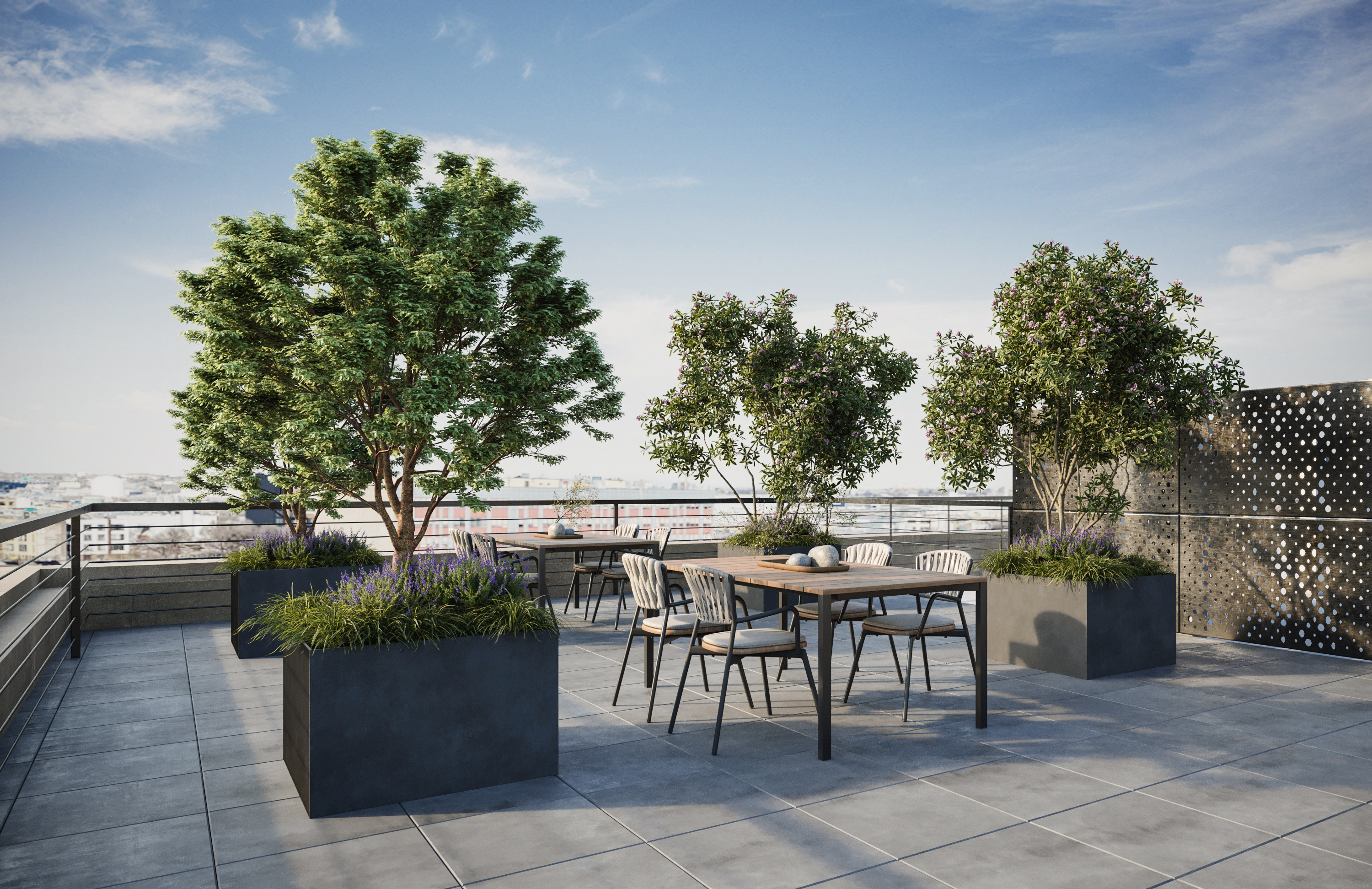 A rooftop terrace with large potted trees, a wooden table and chairs, tiled flooring, and metal railings, with a city skyline visible in the background under a clear sky.