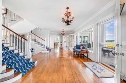 A spacious, sunlit living room with high ceilings, white walls, and large windows that offer sweeping ocean views. The room is furnished with elegant, coastal-inspired décor: plush sofas, a grand fireplace, and nautical accents. Hardwood floors and exposed beams add warmth and character.