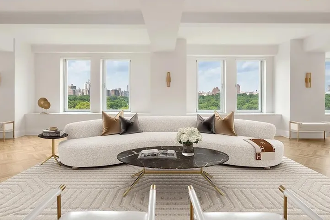 Modern living room with a curved white sofa, brown and gray pillows, a black marble coffee table with a book and flowers, and large windows showing greenery and city buildings, set against light walls and parquet flooring.