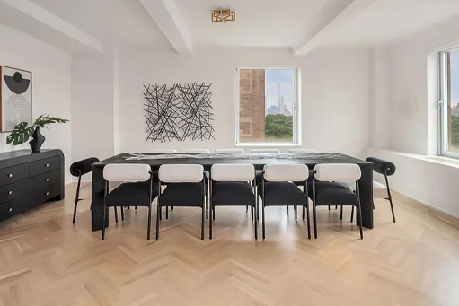 Modern dining room with a long black table, eight black and white chairs, large abstract wall art, two windows letting in natural light, and a black sideboard with a plant.