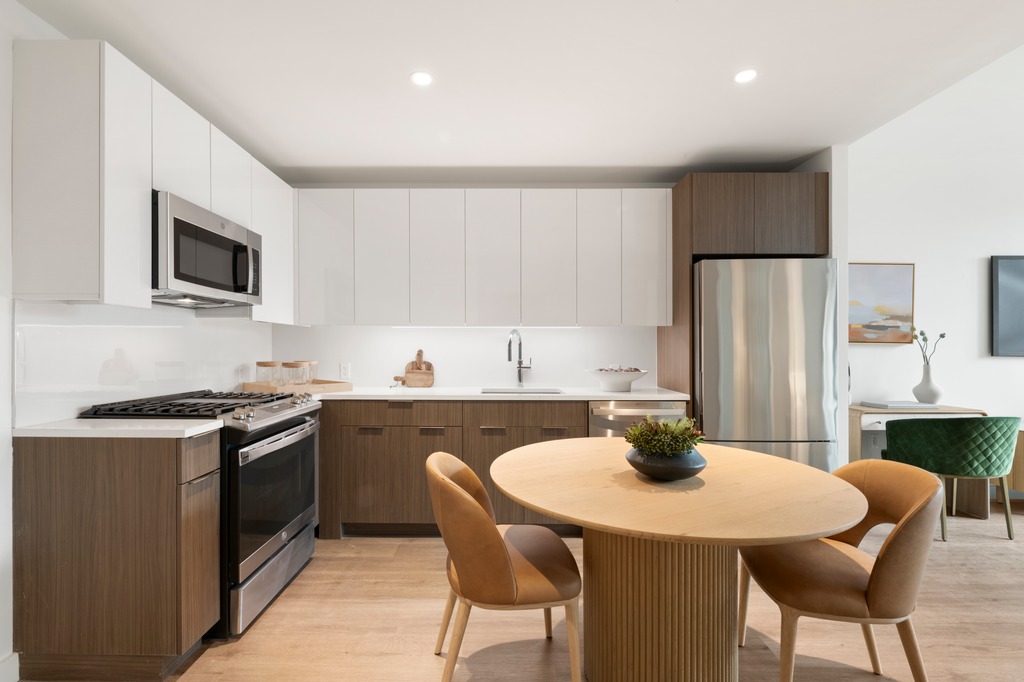 Modern kitchen with stainless steel appliances, white upper cabinets, and wooden lower cabinets. A round dining table with four brown chairs sits in the center, with a small green chair near a desk in the background.