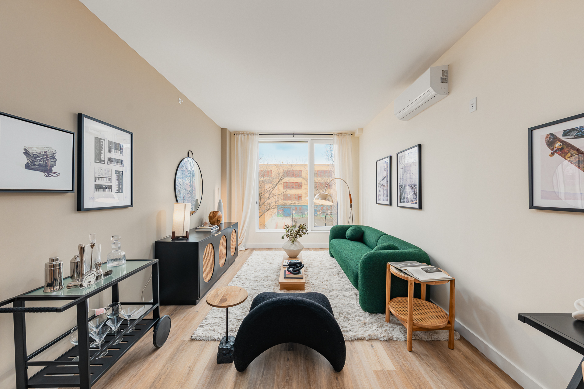 A modern living room with a green sofa, black coffee table, framed artworks on beige walls, a large window with sheer curtains, a light-colored rug, and wooden flooring.