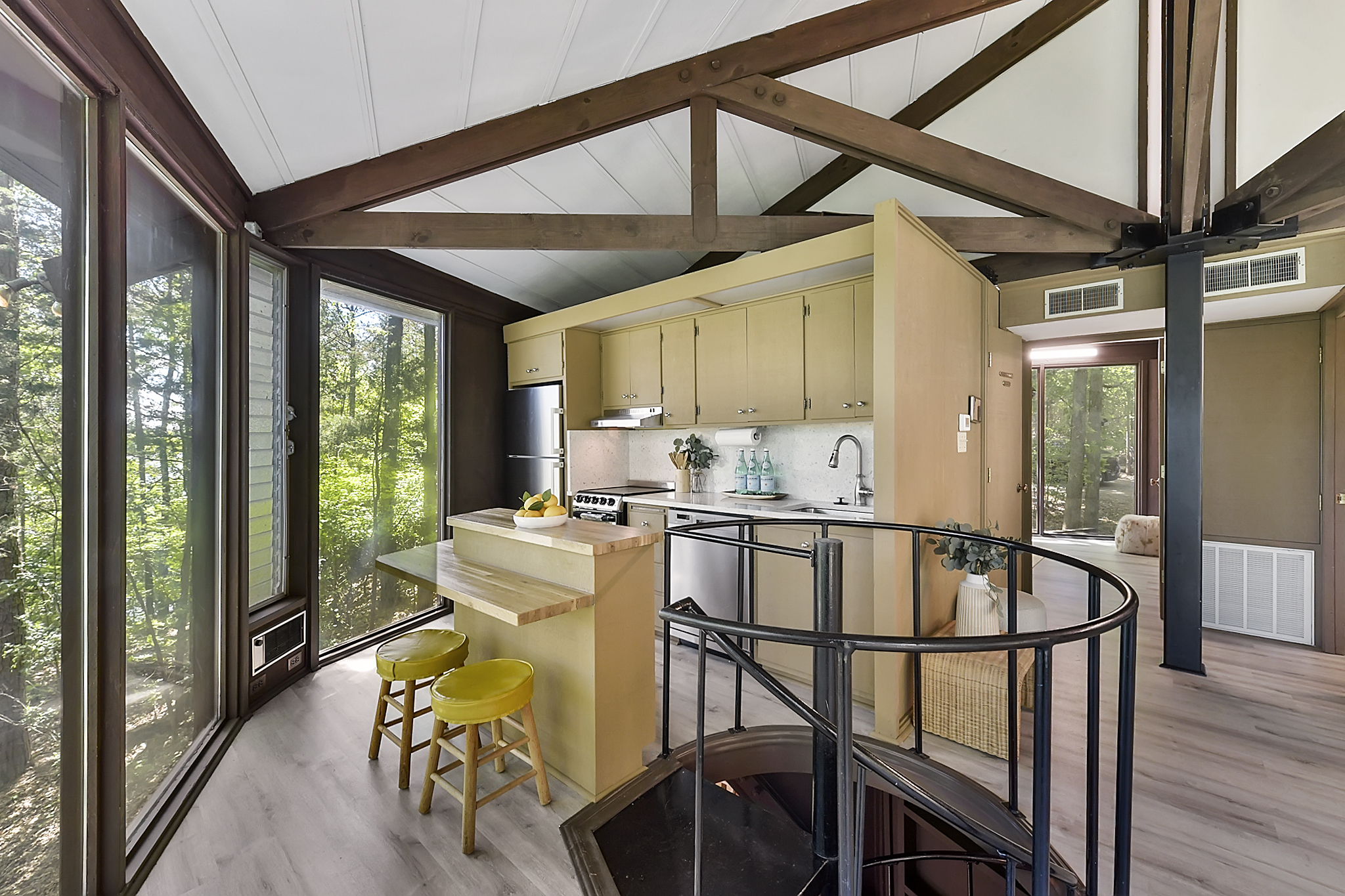 A modern kitchen with light wood cabinets, a small island with two yellow stools, and a spiral staircase leading down. Large windows provide a view of the surrounding forest. Exposed wooden beams are visible on the ceiling.
